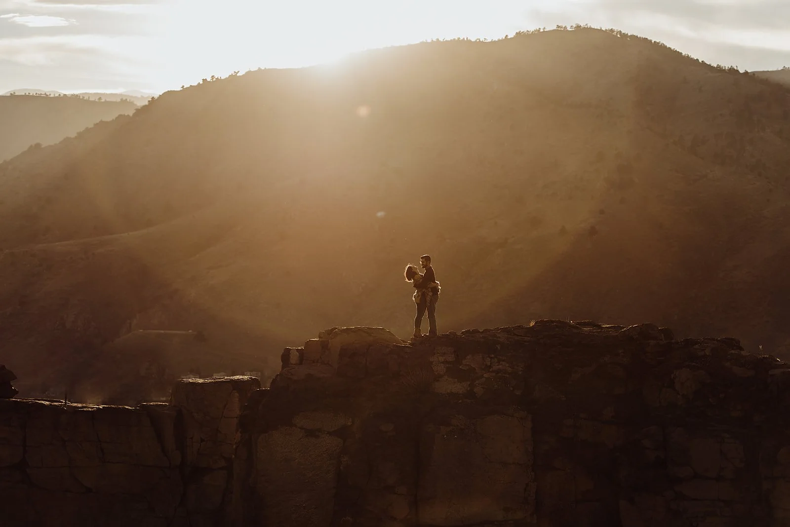 A couple embraces on a rocky cliff at sunset with mountains in the background.
