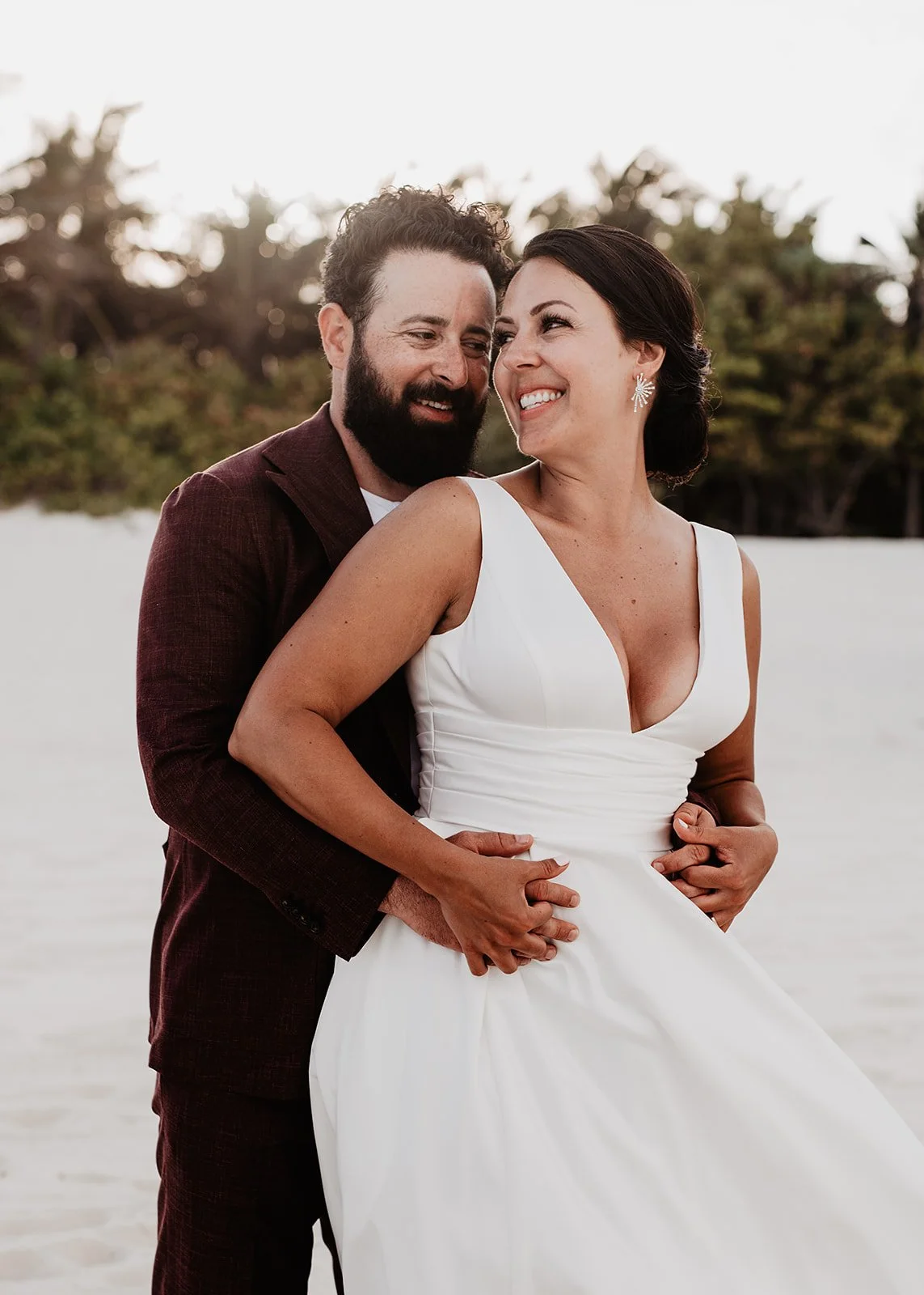 A couple embracing on a beach with trees in the background; the woman is in a white dress, and the man is wearing a suit.