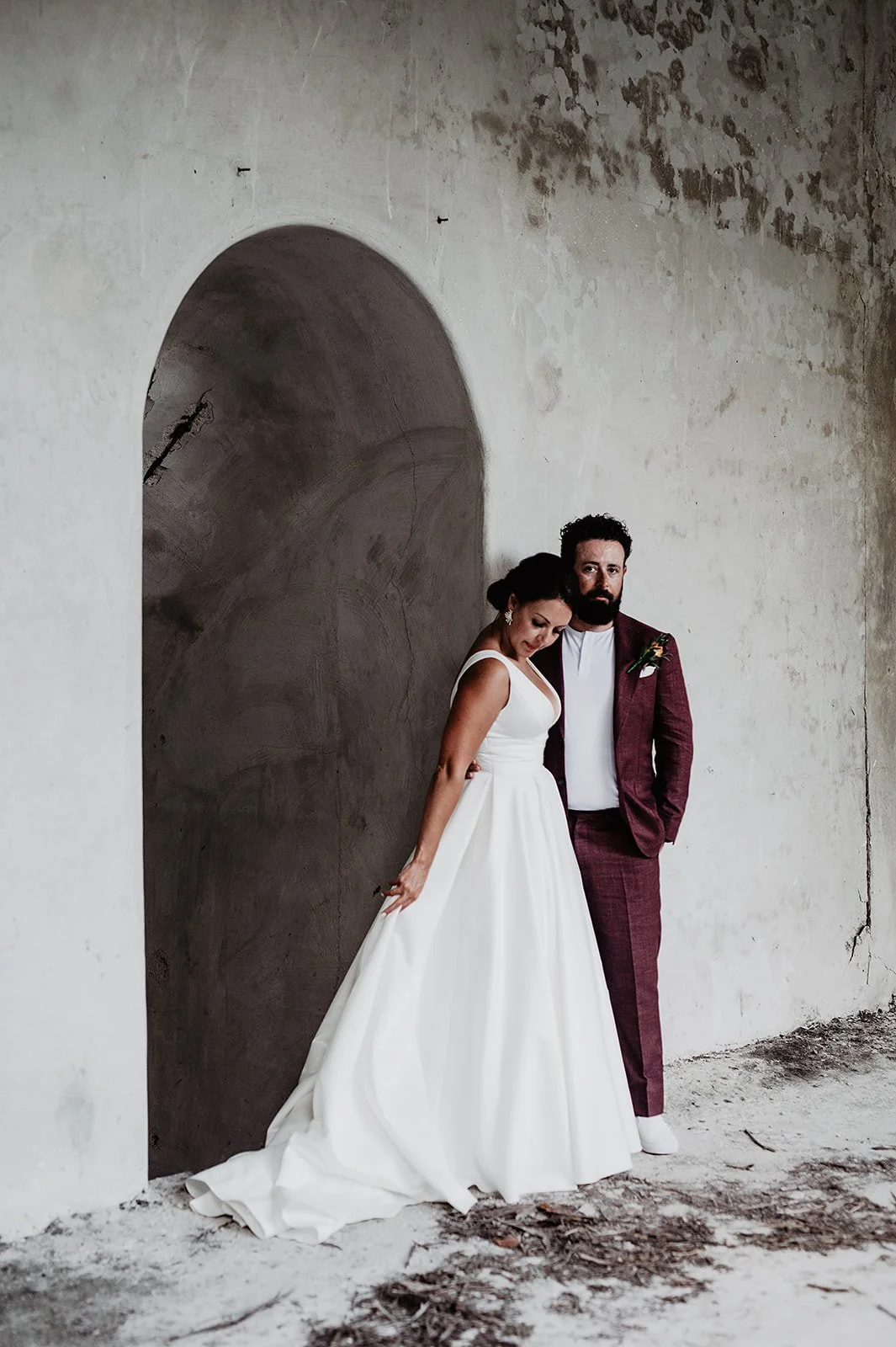 A bride in a white gown and a groom in a maroon suit standing against a textured wall.