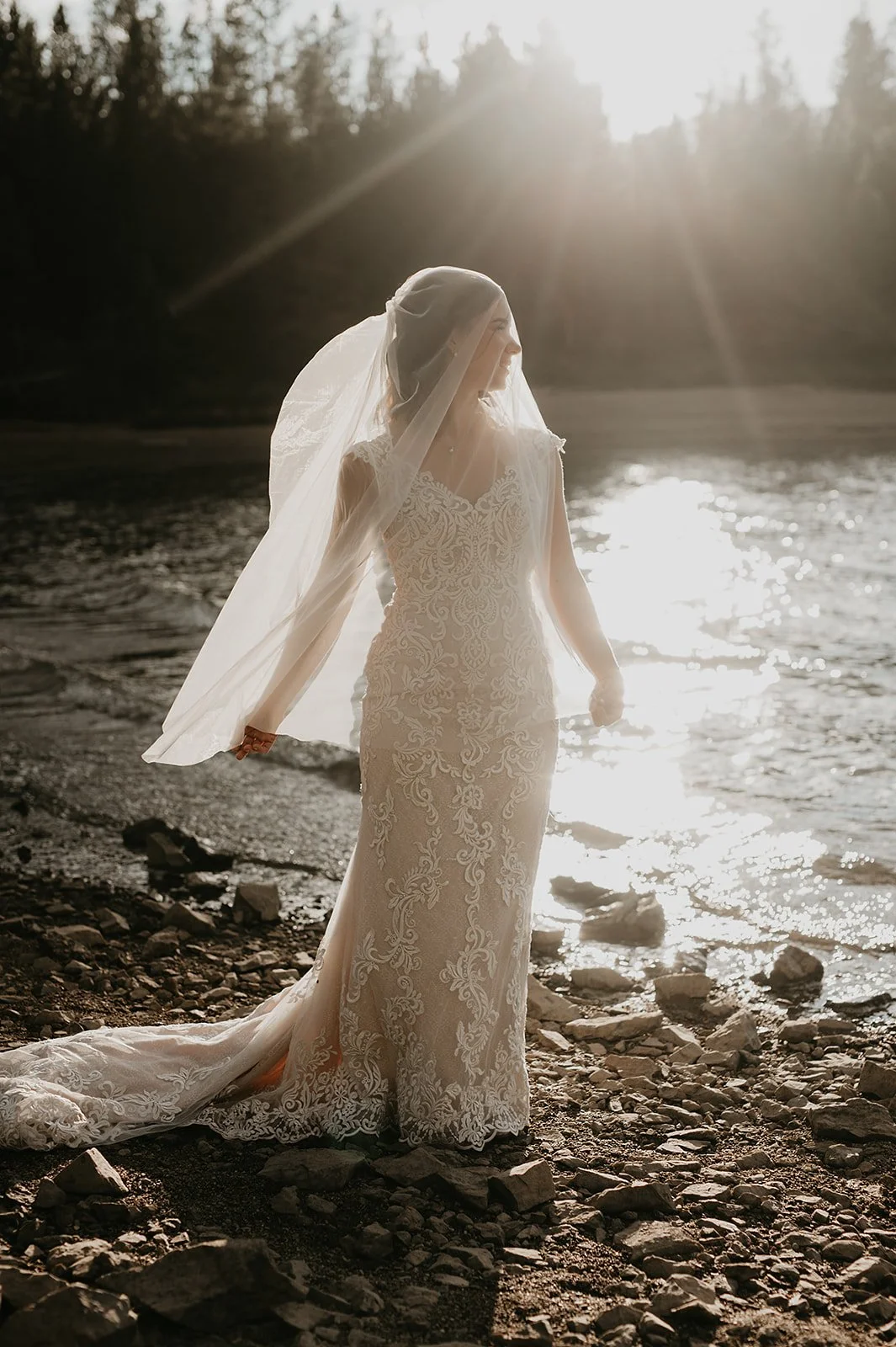 Bride in lace wedding dress with veil standing by a sunlit riverbank.