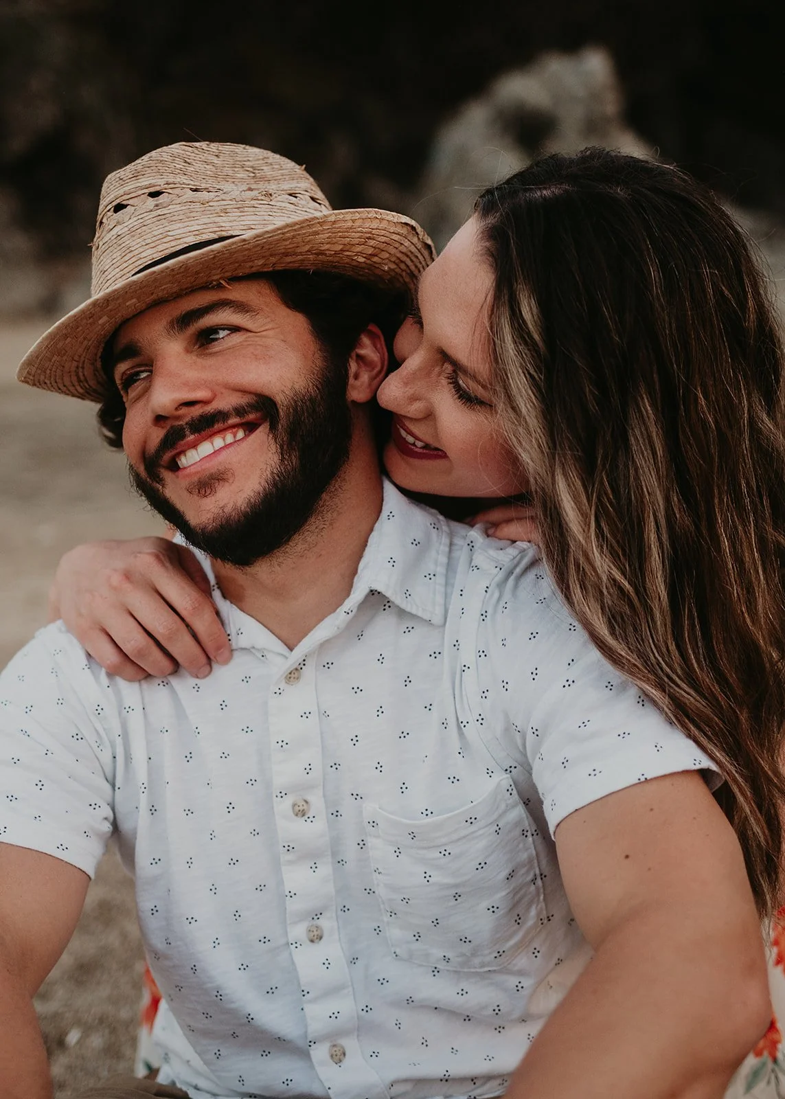 A smiling man in a straw hat and white shirt is sitting with a woman embracing him from behind. They appear happy and relaxed in an outdoor setting.