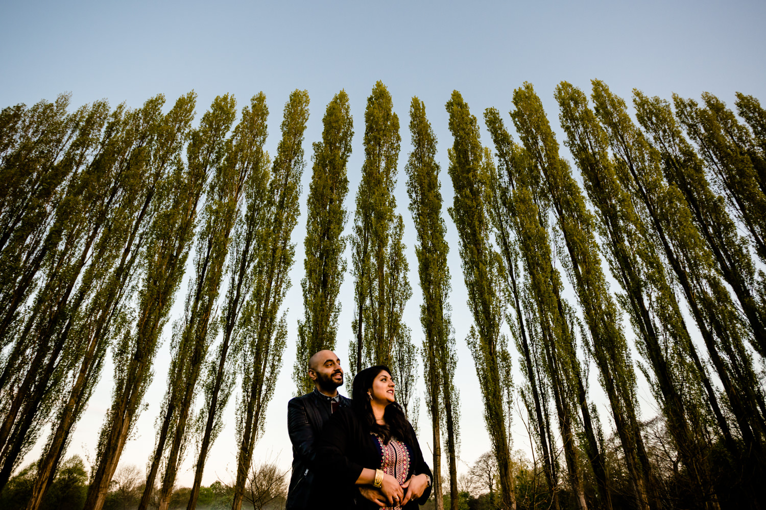 Hugging couple in front of a row of tall trees on their engagement shoot in a Manchester park.