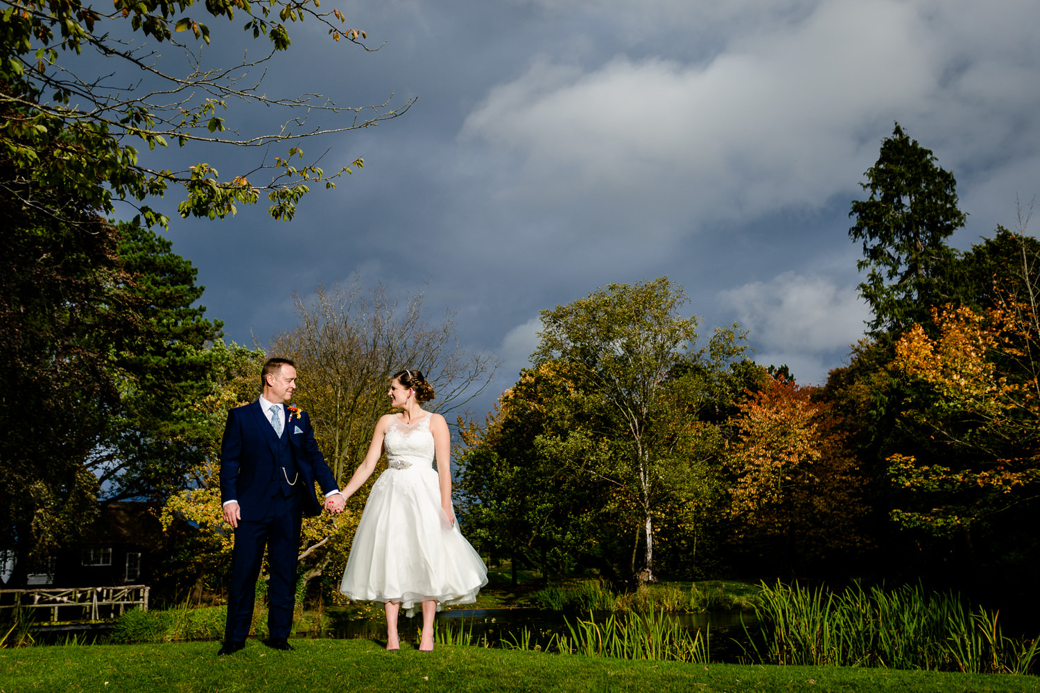 A bride and groom holding hands stood by a lake in front of autumn trees at Thornton Manor in Wirral