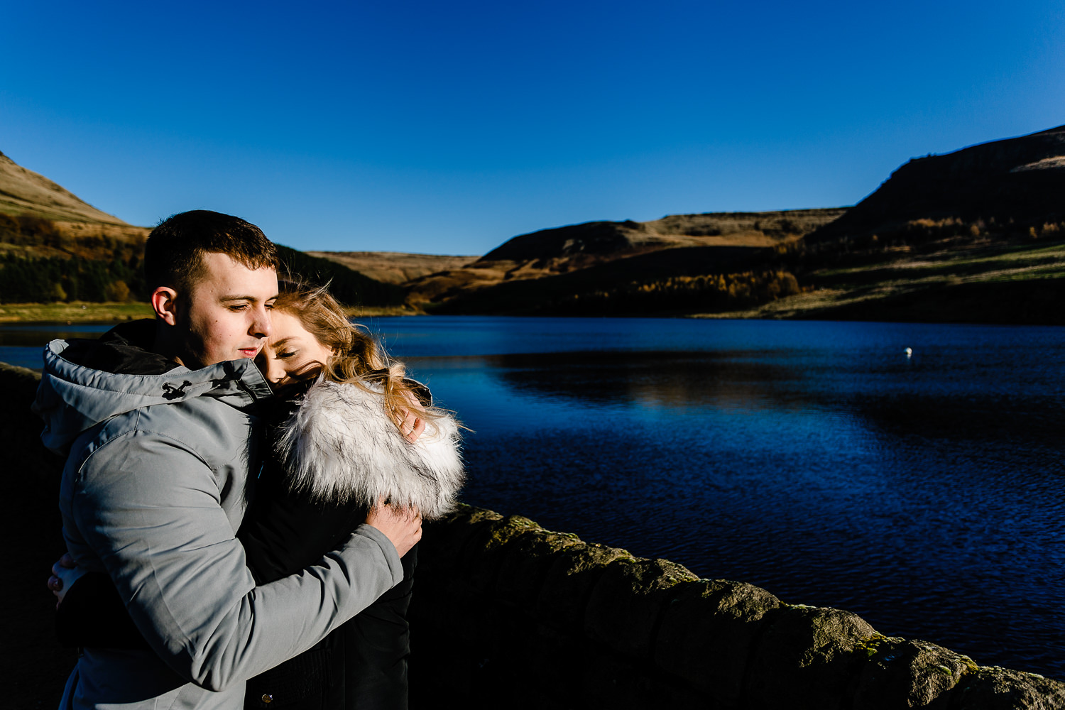 An engaged couple keep each other warm beside the deep blue Dovestone reservoir, pre wedding shoot by Manchester wedding photographers.