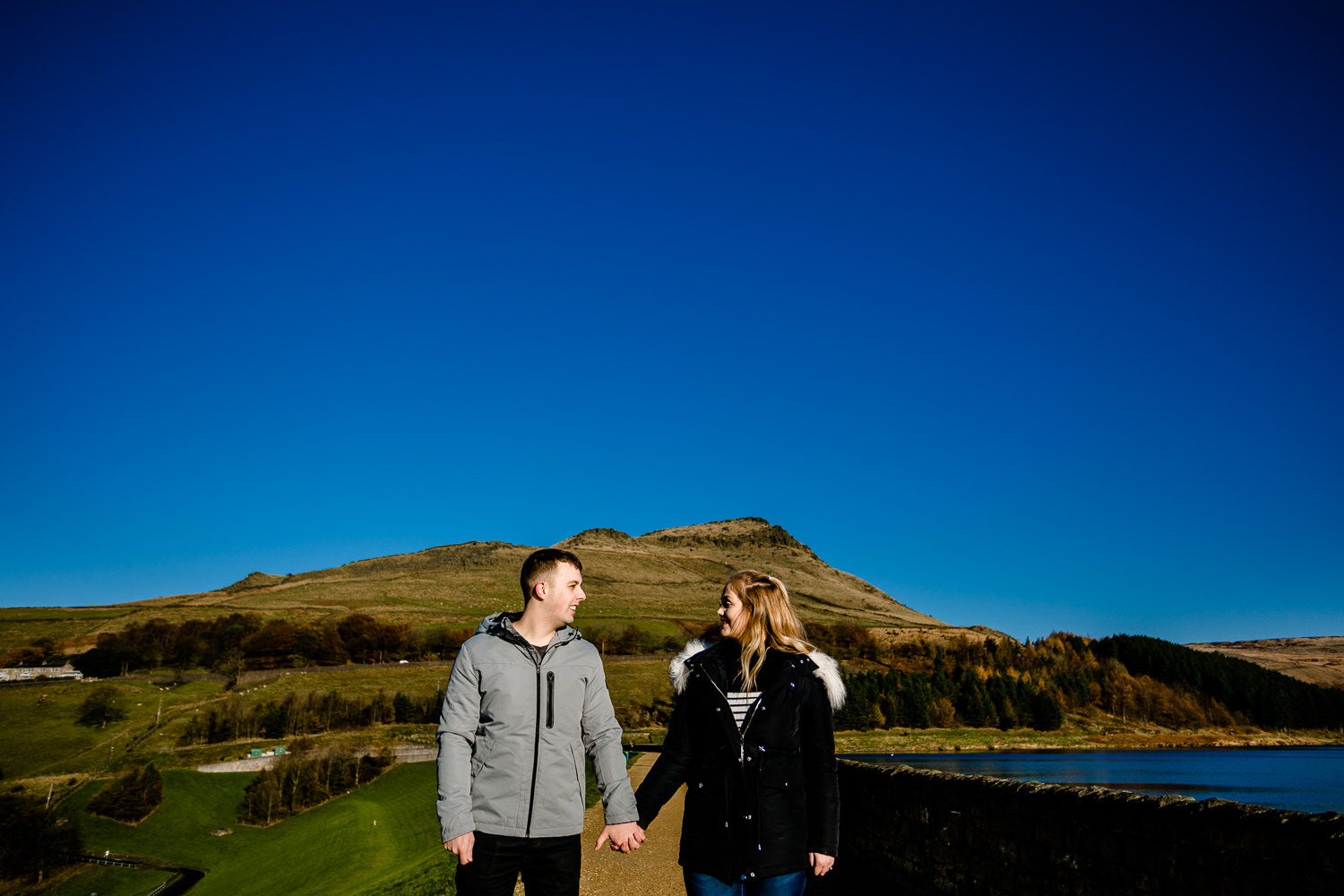 Sarah and Jason stand in front of a beautiful landscape on a sunny Manchester winters day on their Dovestone Reservoir pre wedding shoot.