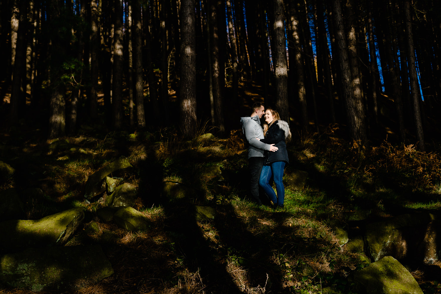 A couple stood in dappled light under pine trees. 