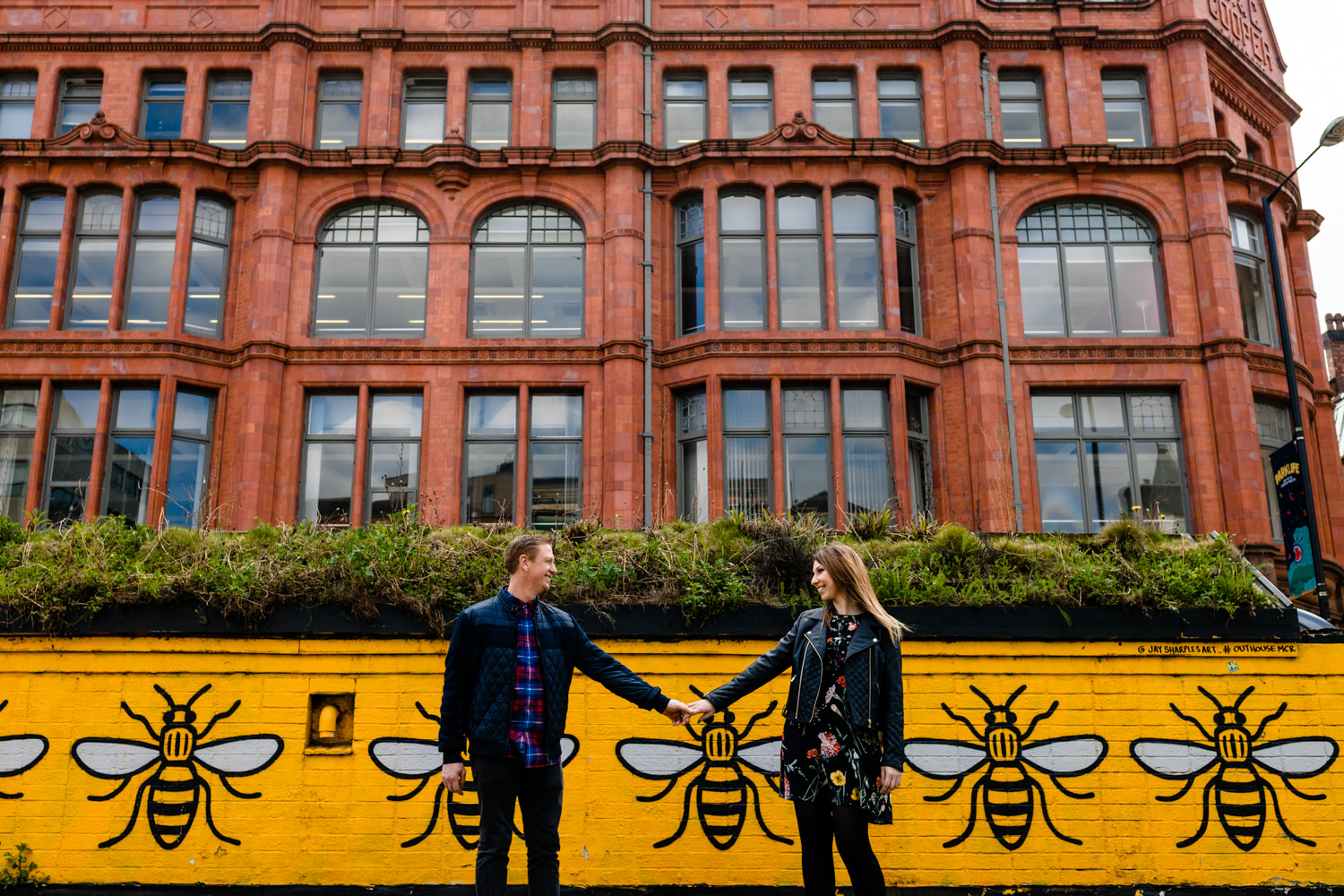 Urban pre wedding shoot in Manchester's Norther Quarter, a couple hold hands stood in front of a wall mural of the iconic yellow Manchester bees. 