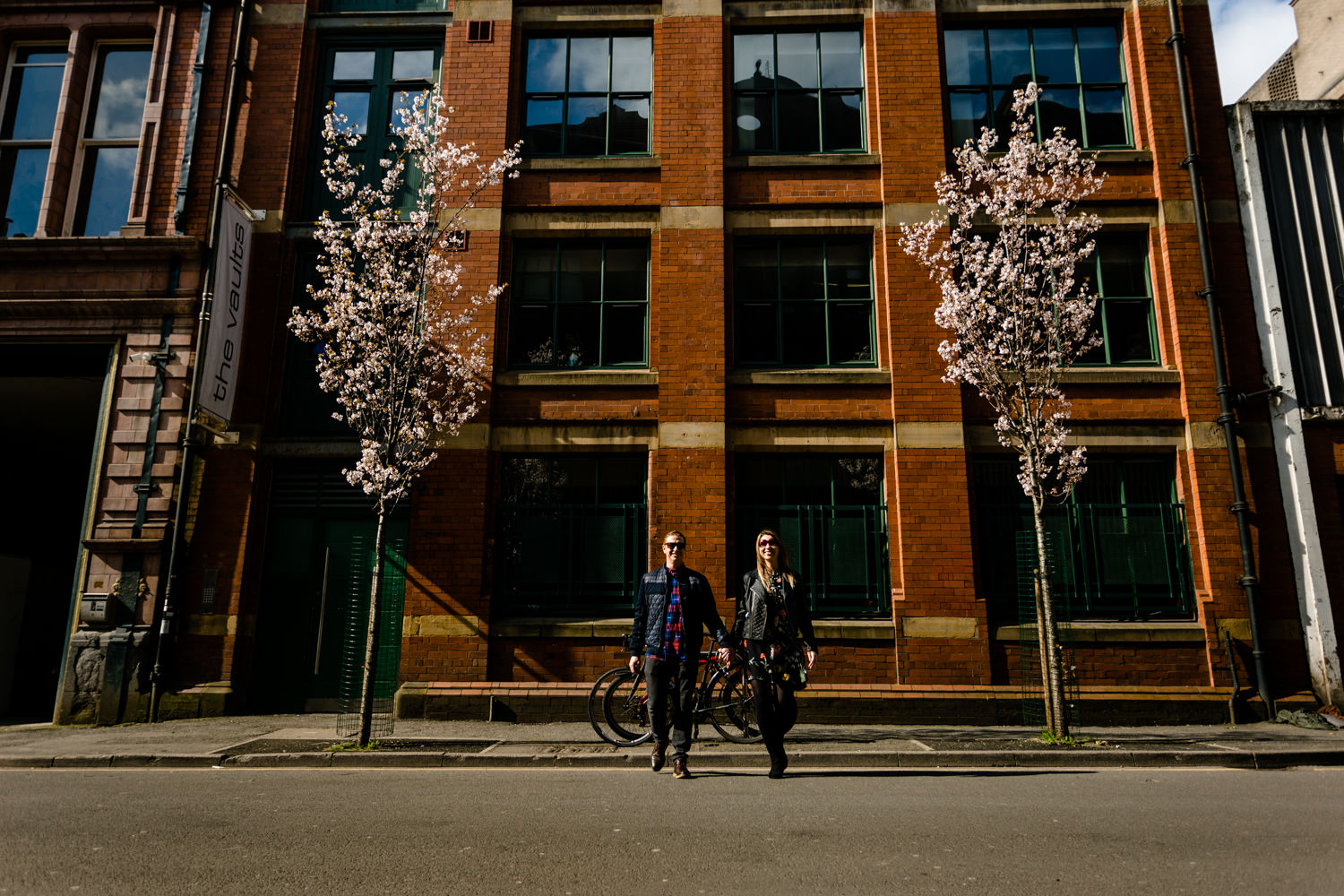 A couple cross the street in the Northern Quart of Manchester between some blossom trees. 