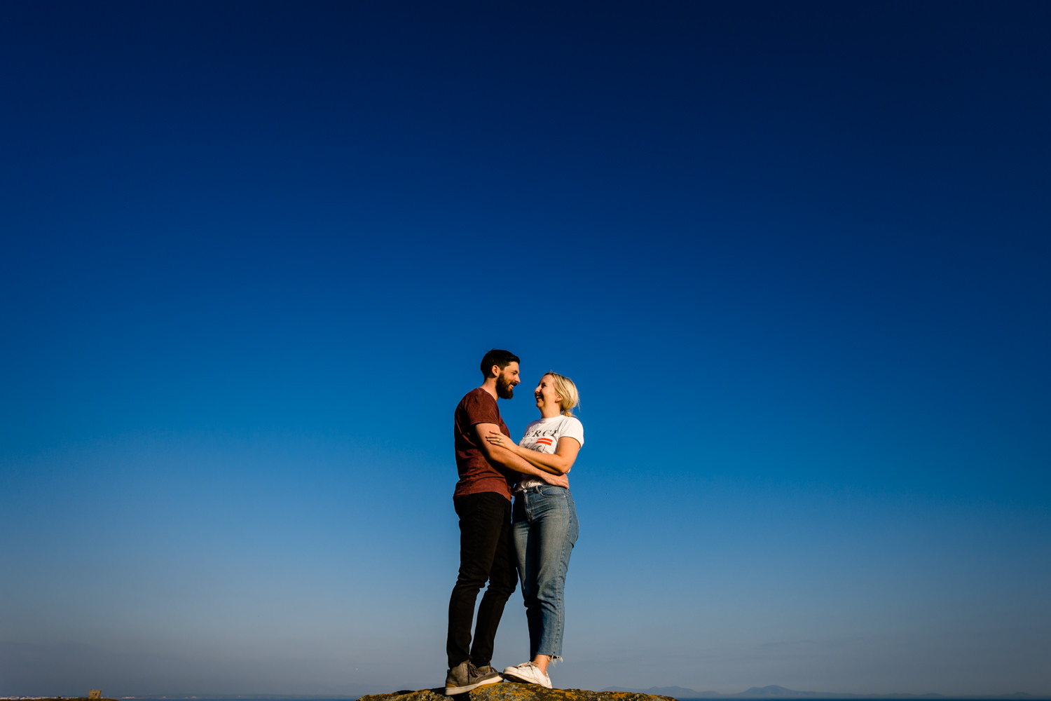 An engaged couple stood on a cliff in front of a blue sunny sky in Anglesey, Wales. By wedding photographers Zoe & Tom of About Today Photography.