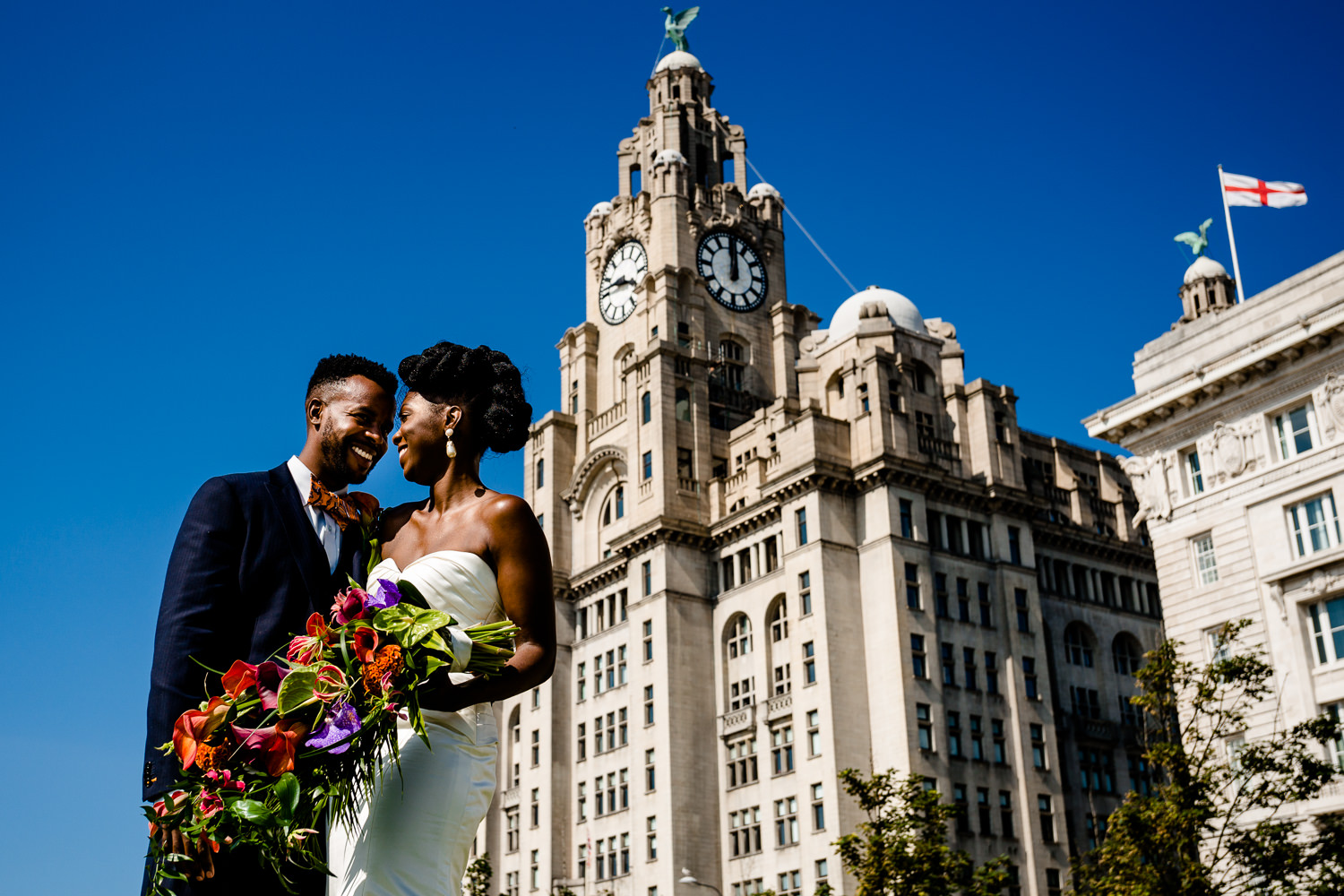 Bride & Groom with a colourful, tropical bouquet standing and smiling at each other in front of the Liver Building, by Liverpool wedding photographers.