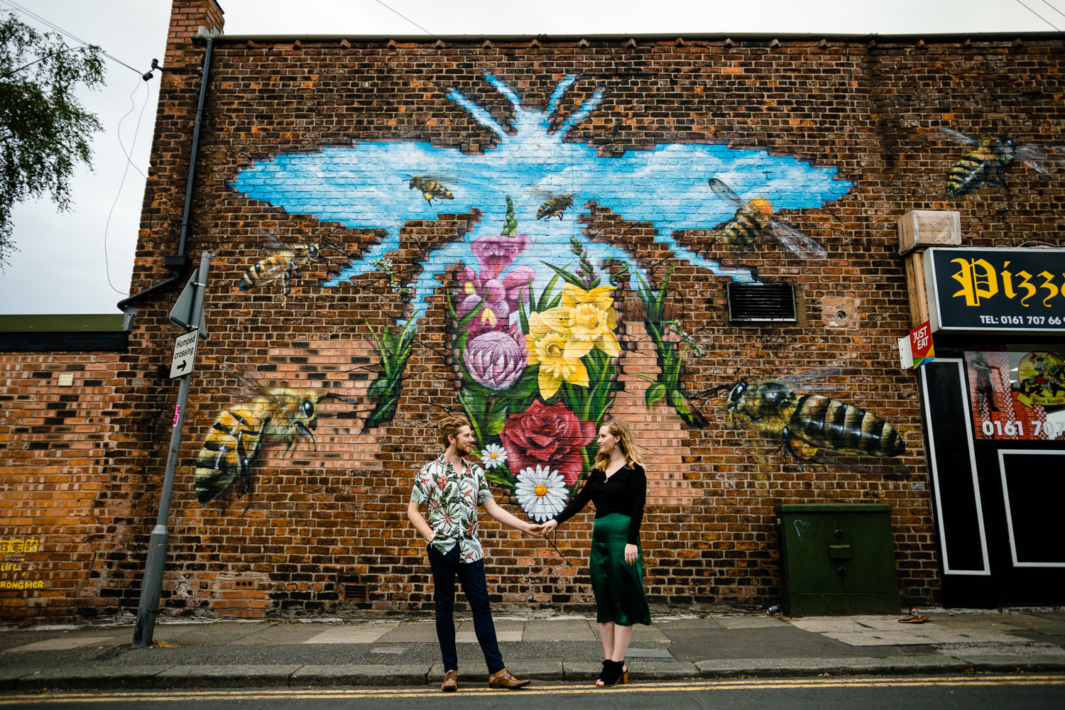 Rose & Josh stood in front of a colourful Manchester Bee wall mural in Monton, by colourful wedding photographers in Manchester.