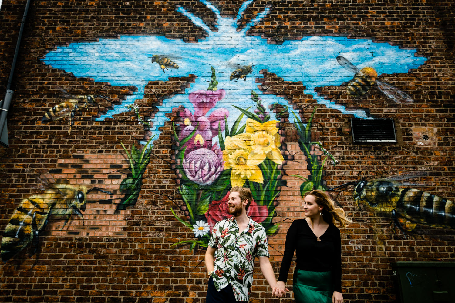 Colourful pre wedding shoot, a couple holding hands in front of a Manchester Bee ahead of their wedding day in Chorlton.