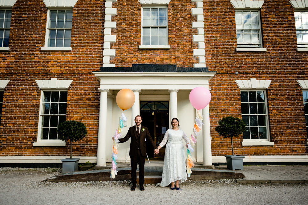A bride and groom holding giant balloons with colourful tassels, at their Chester wedding outside a hall.