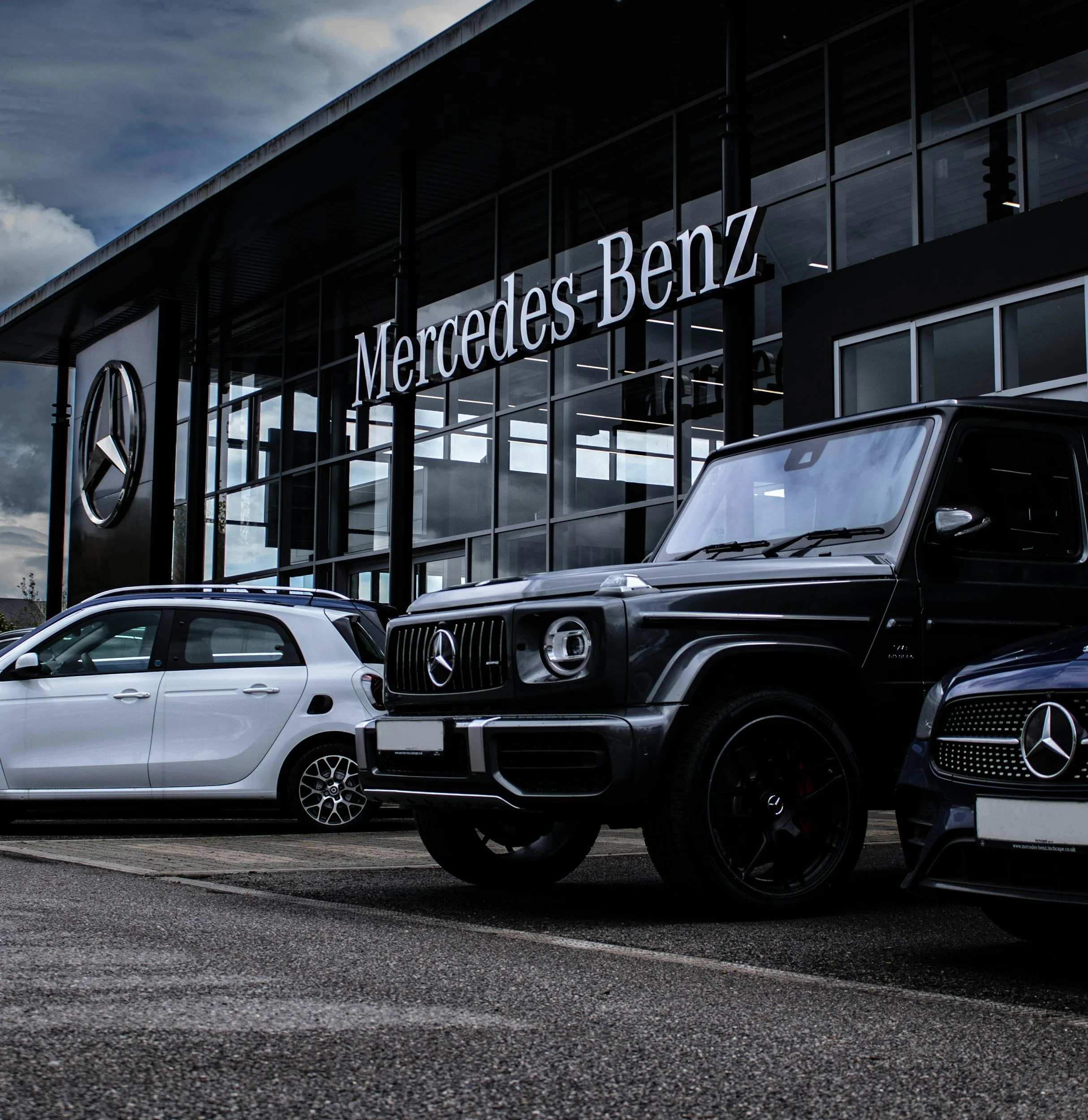 Mercedes-Benz dealership with three Mercedes vehicles parked in front, one silver smart car and two black larger luxury SUVs, alongside a modern glass-front showroom with the Mercedes-Benz logo and name.