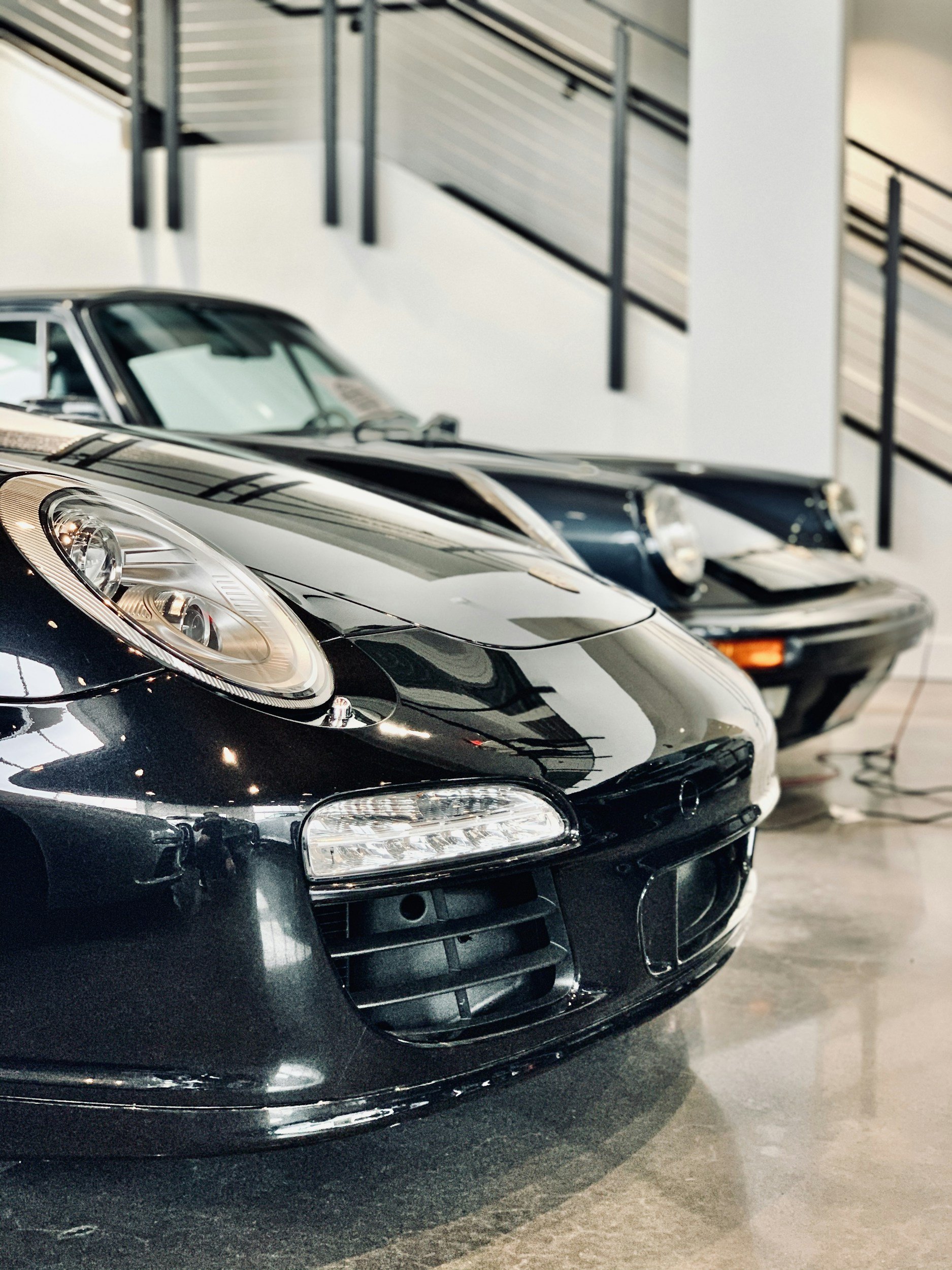 Close-up of two classic sports cars, black in the foreground and dark blue in the background, in an indoor showroom with modern decor and railing.