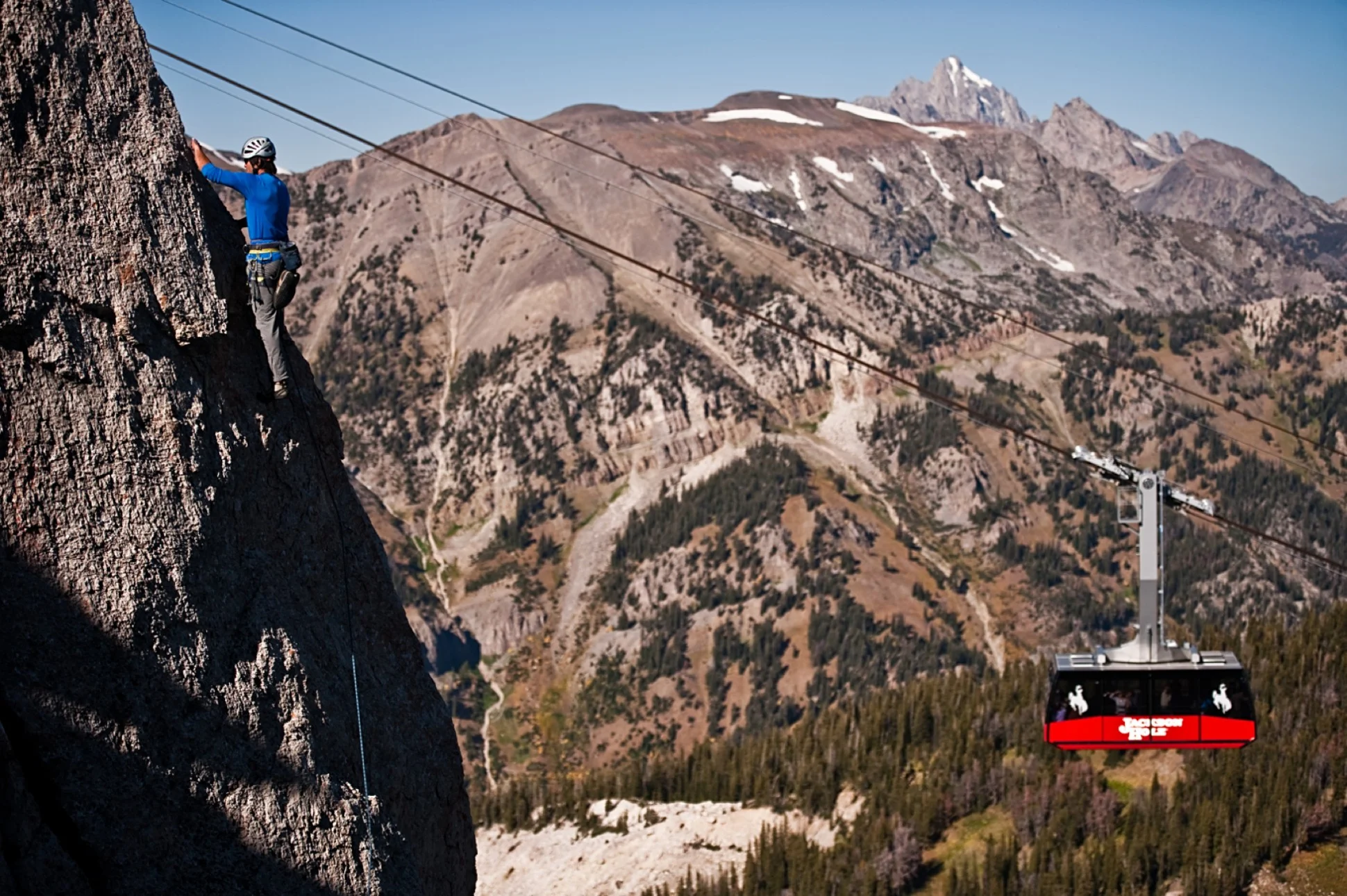 A rock climber makes his way up a face as a Jackson Hole tram car moves behind him