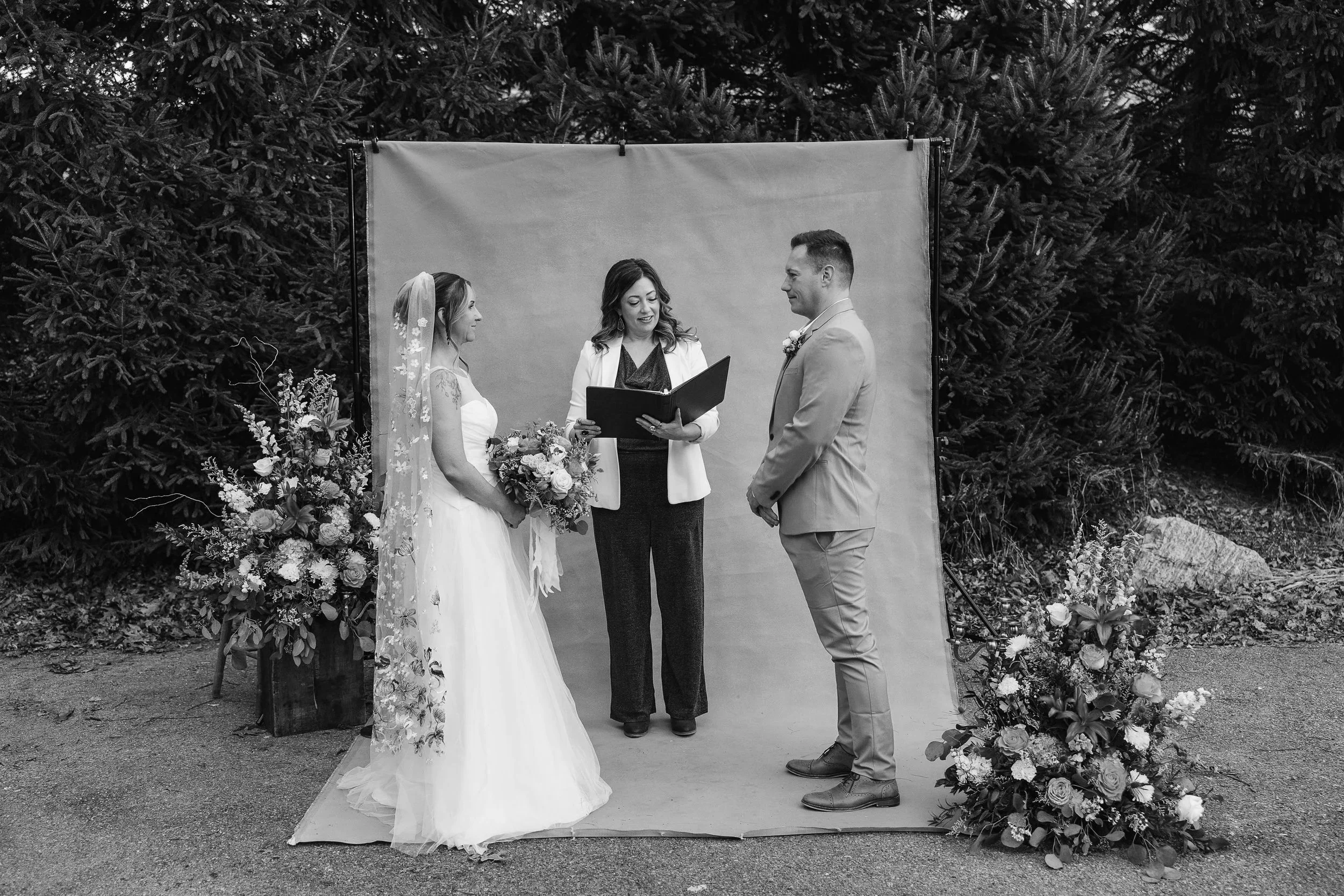 A black-and-white photo of a wedding ceremony outdoors with a backdrop of trees, featuring a bride in a wedding gown and veil, a groom in a suit, and a officiant holding a book.