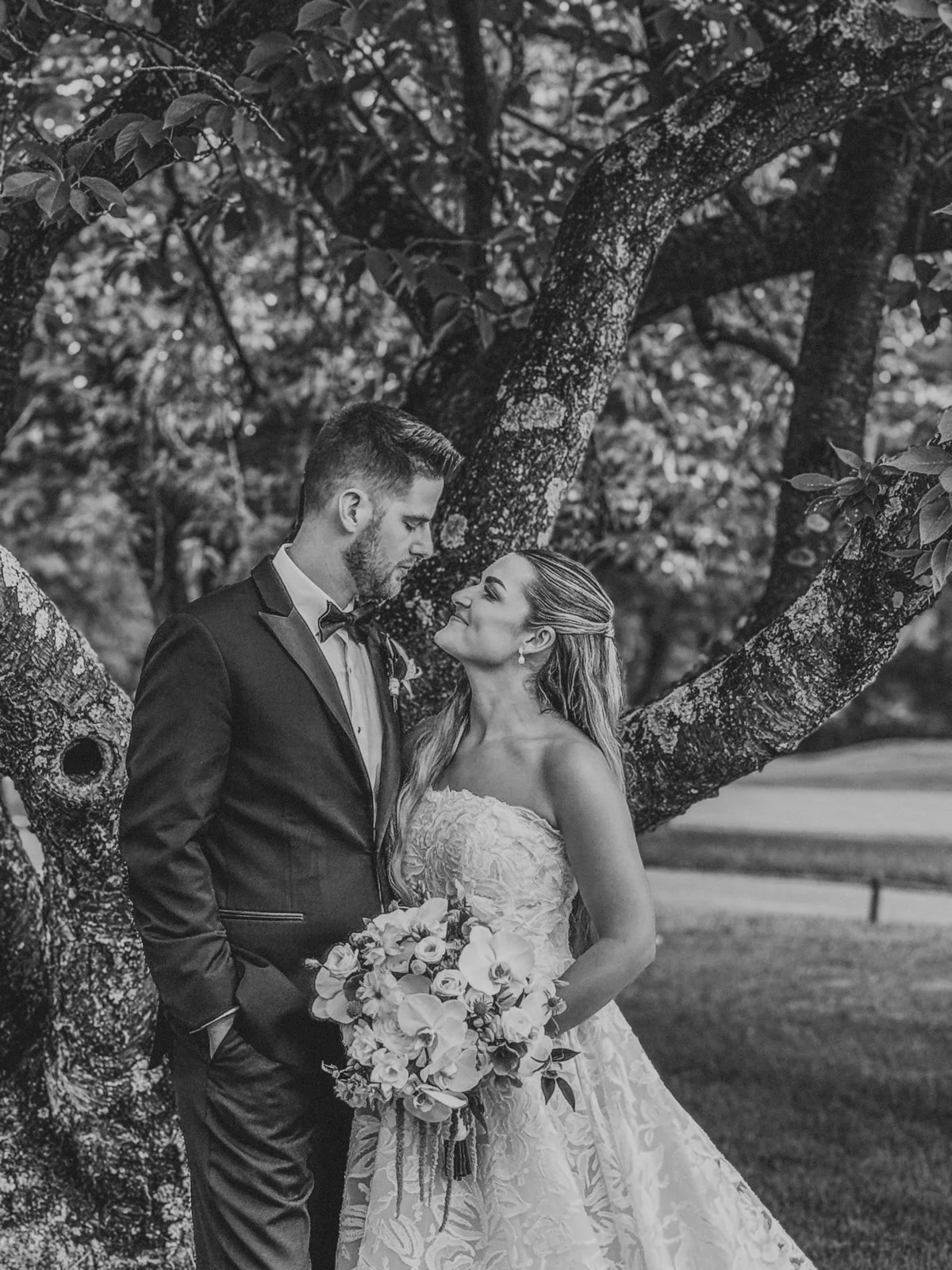 Black and white photo of a wedding couple standing close to each other outdoors, with a large tree behind them. The groom is wearing a tuxedo and the bride is in a strapless wedding gown, holding a bouquet of flowers, and looking into each other's eyes.