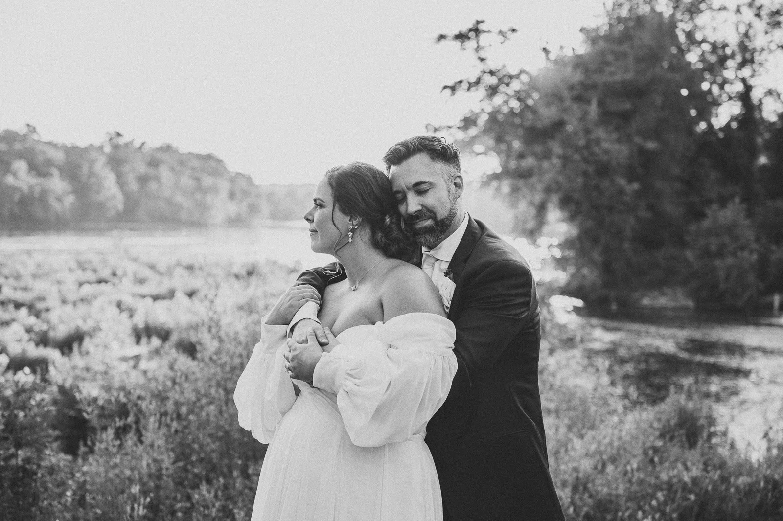 Black and white photo of a bride and groom embracing in a natural outdoor setting, with the bride in an off-shoulder wedding dress and the groom in a suit.