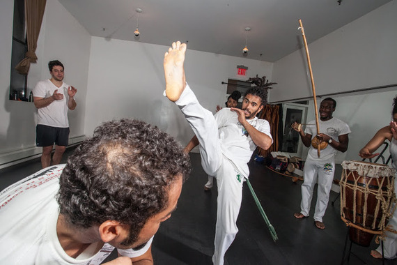 Students learn dozens of basic kicks, ducks and maneuvers which can be assembled in infinite variation, and practice them in a game of constant motion and surprise, as seen above with Pena kicking a Bênção and a student practicing a duck, or Esquiva, all done to the type of game determined by the music. (Photo by Ace Murray / Brooklyn Juice Photography .