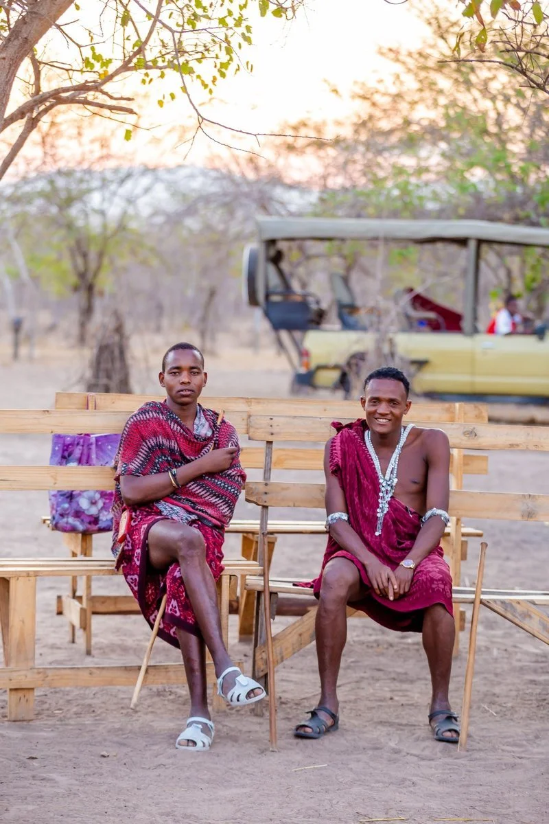 Two Maasai men sit outdoors on safari with a safari vehicle in the background