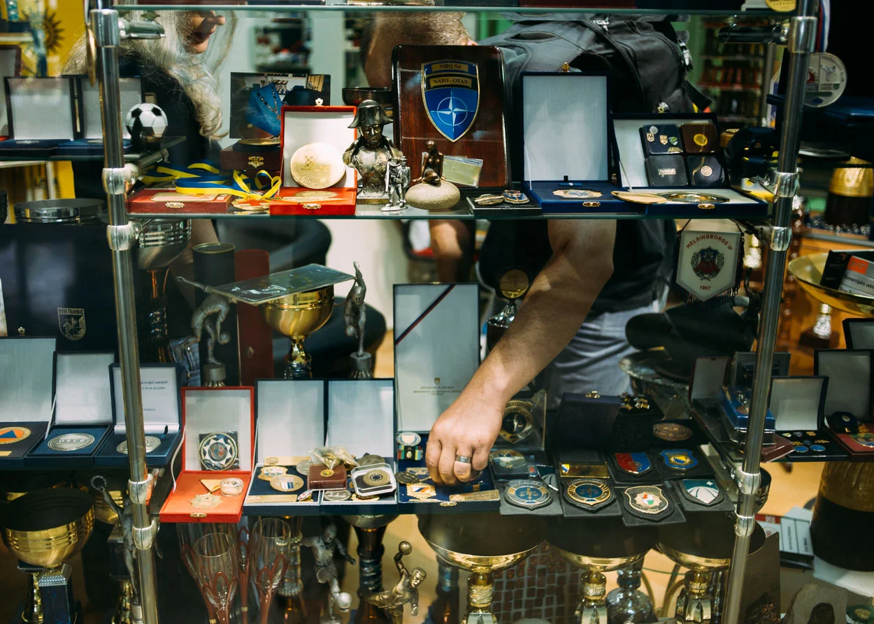 Andrej inspects replica medals at a trophy shop. Also shown in the window are replica police badges, similar to the one on Andrej's hip.