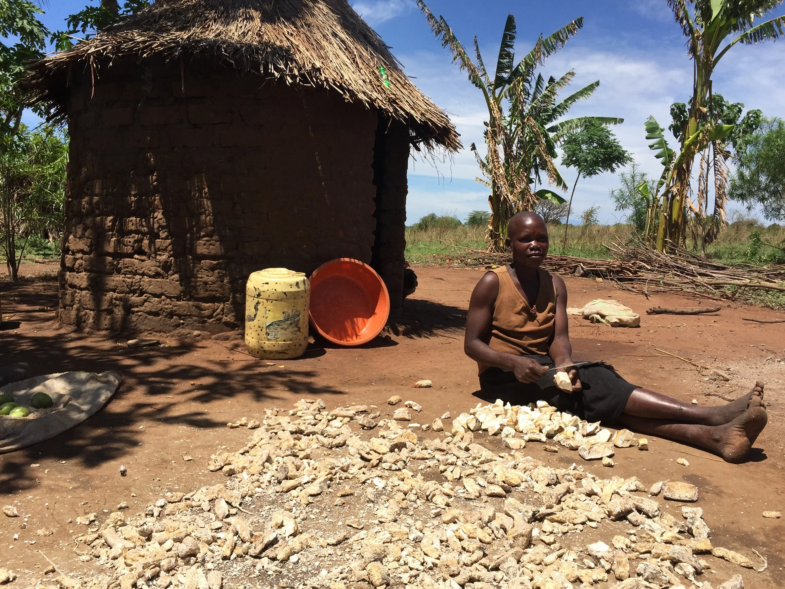 An orphan in front of her home processing food she's grown. She's looking forward to her new home soon.