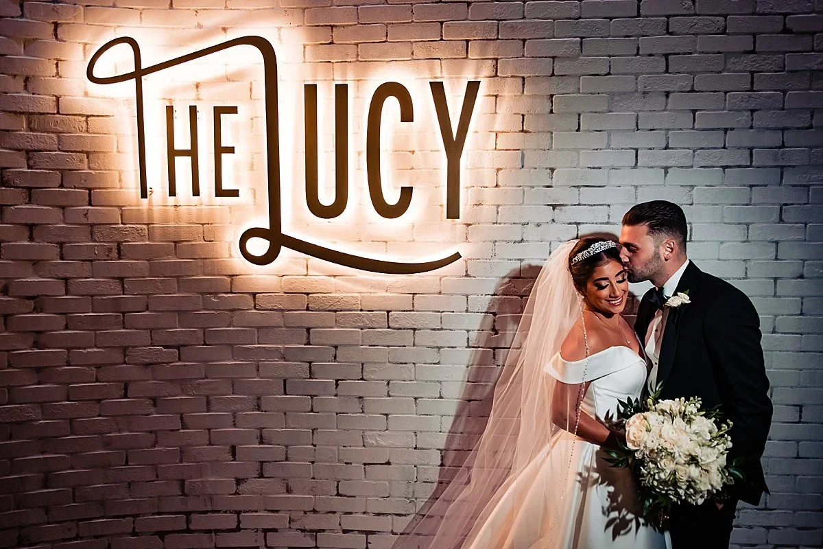 Couple in front of the glowing Lucy sign on the white brick wall