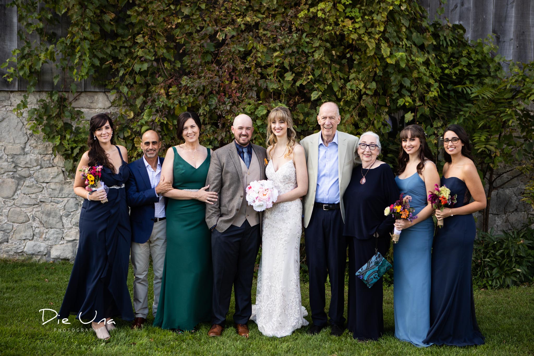 bride and groom with brides family for traditional wedding portrait in front of a barn with plants growing on it.jpg