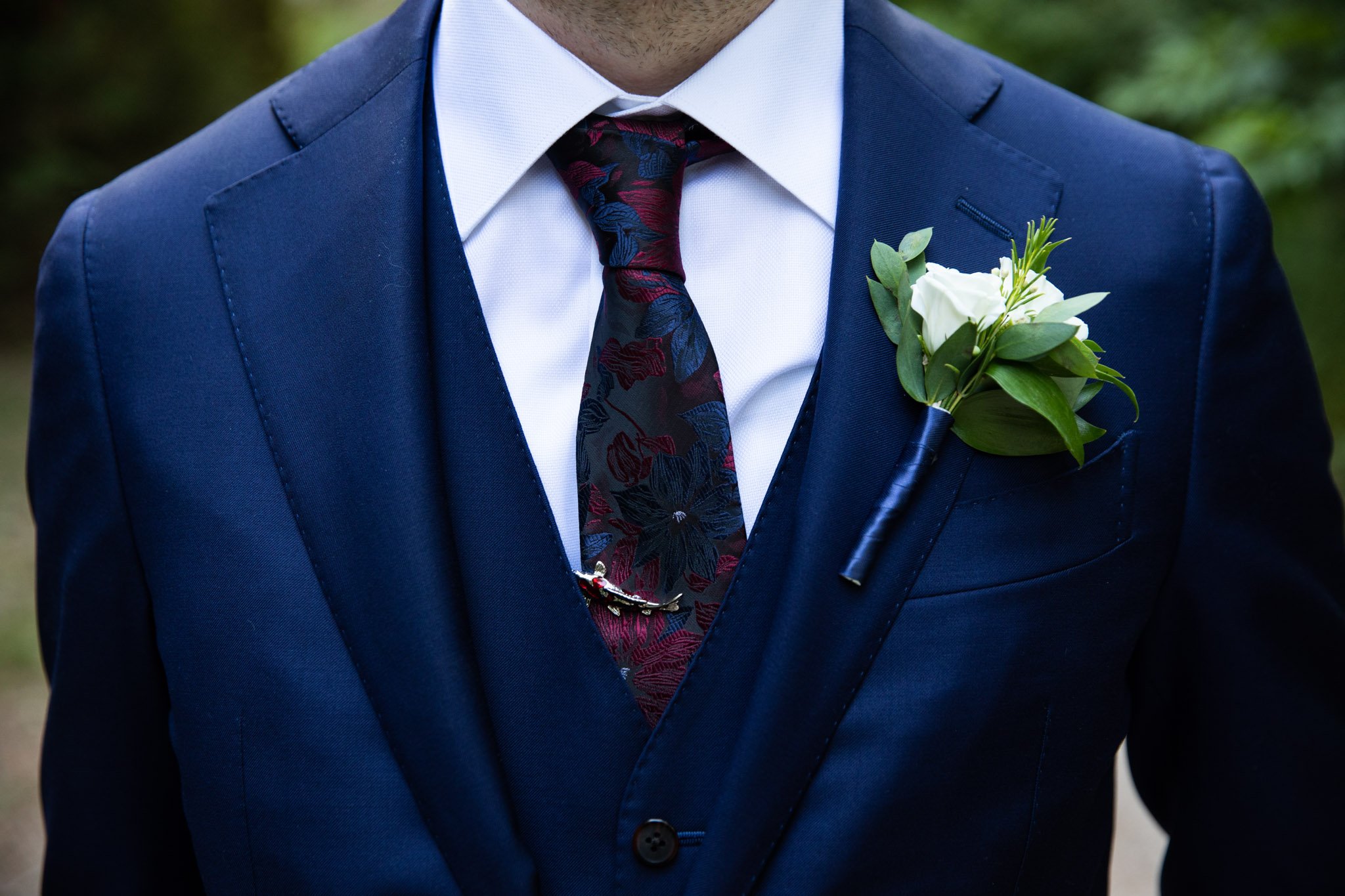 close up of grooms navy suit with vest, tie, tie clip and boutonniere.jpg