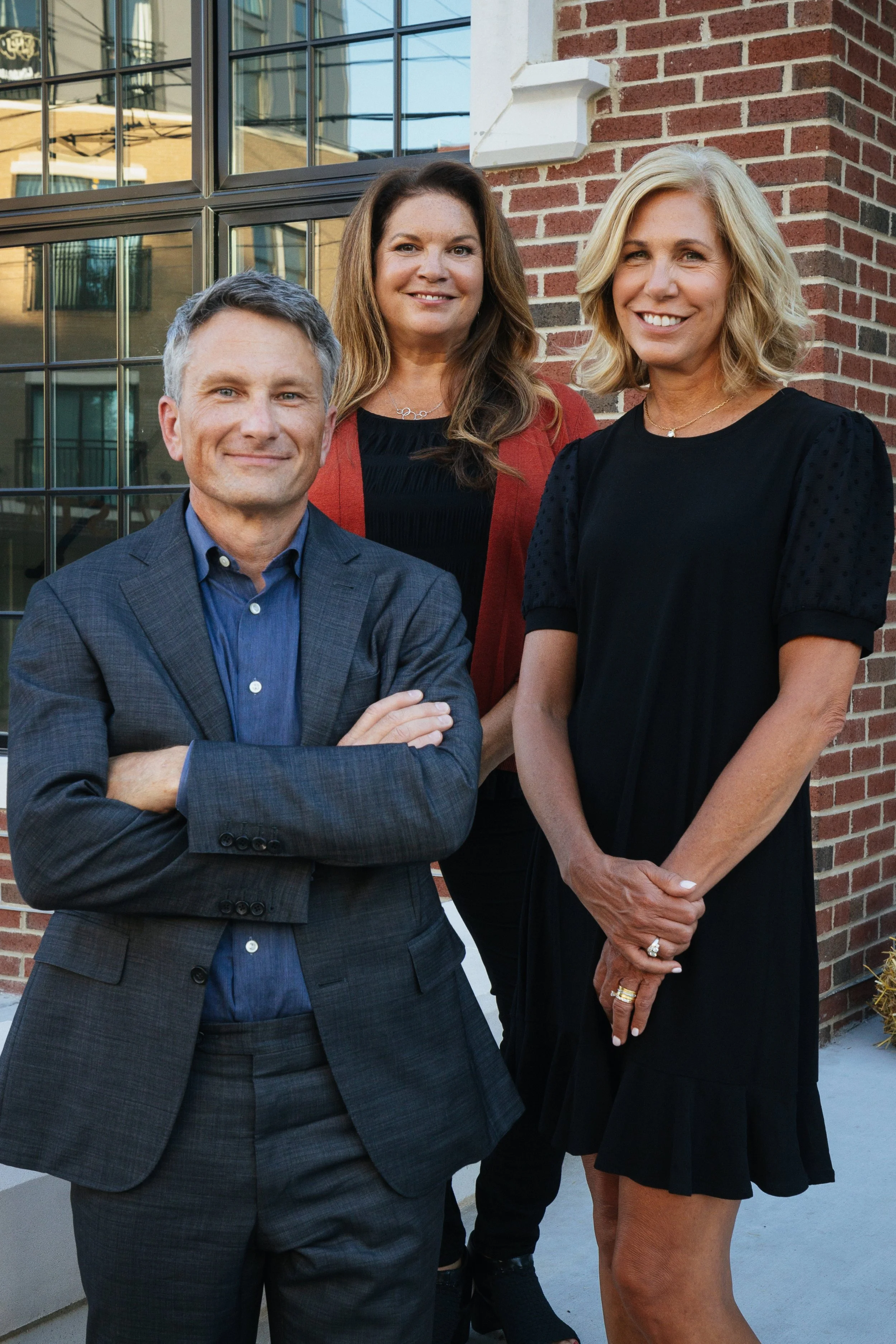 Three adults standing outside in front of a brick building with large windows. The man in front wears a blue shirt and gray suit with arms crossed, smiling. The woman behind him has long brown hair, wears a black dress with a red cardigan, and is smiling. The woman on the right has blonde hair, wears a black dress, and is smiling with her arms relaxed. All are dressed professionally.