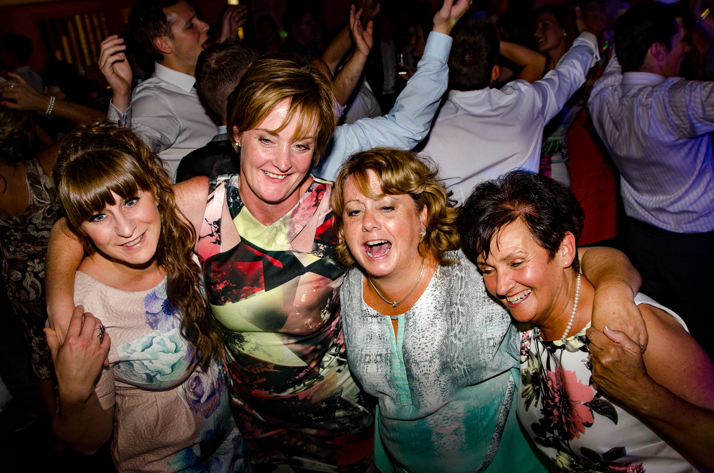 Foreground: Family group on dancefloor enjoying the wedding. Background: More people dancing with arms in the air.