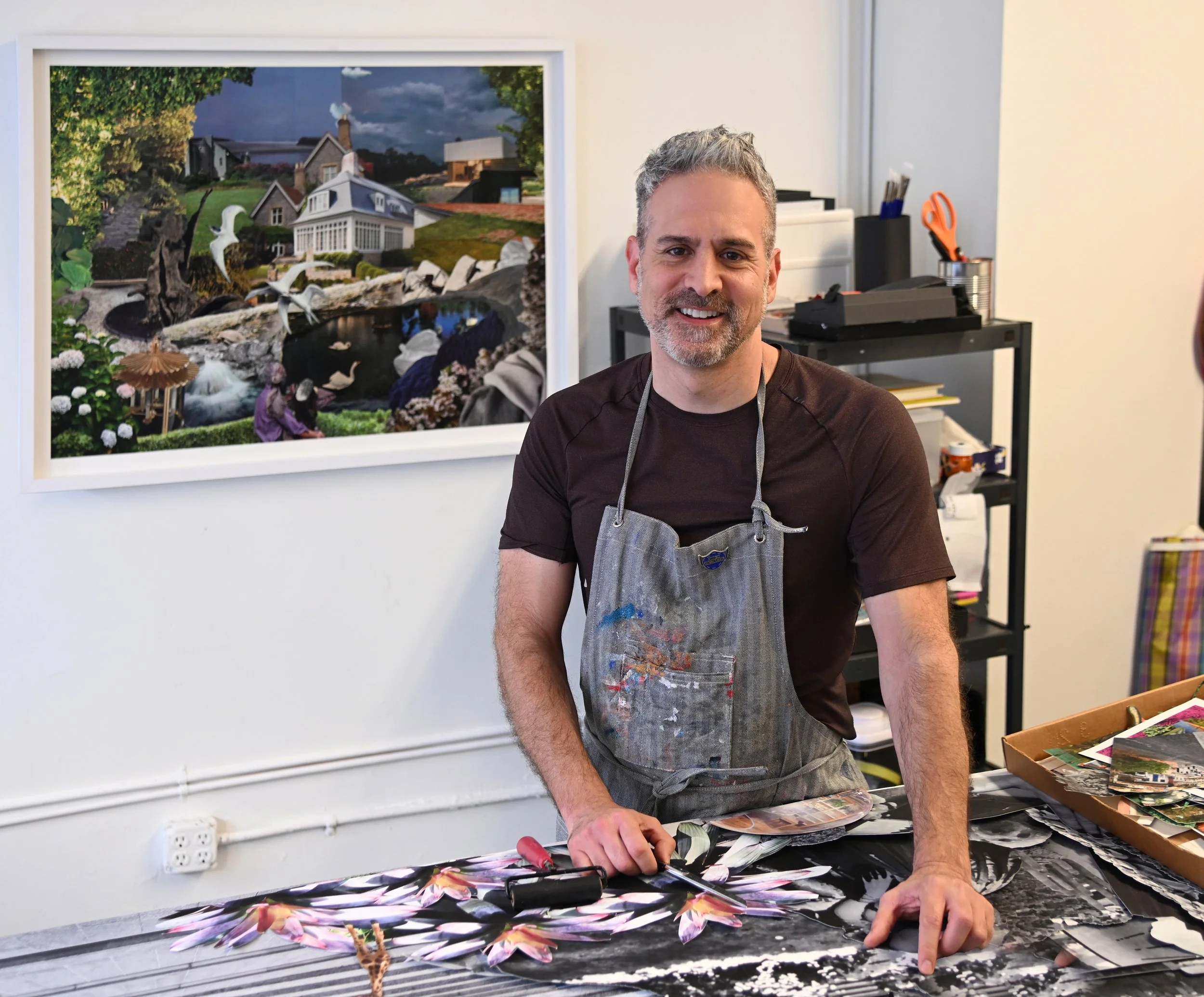 Artist in his studio, wearing apron, standing over worktable with collage materials