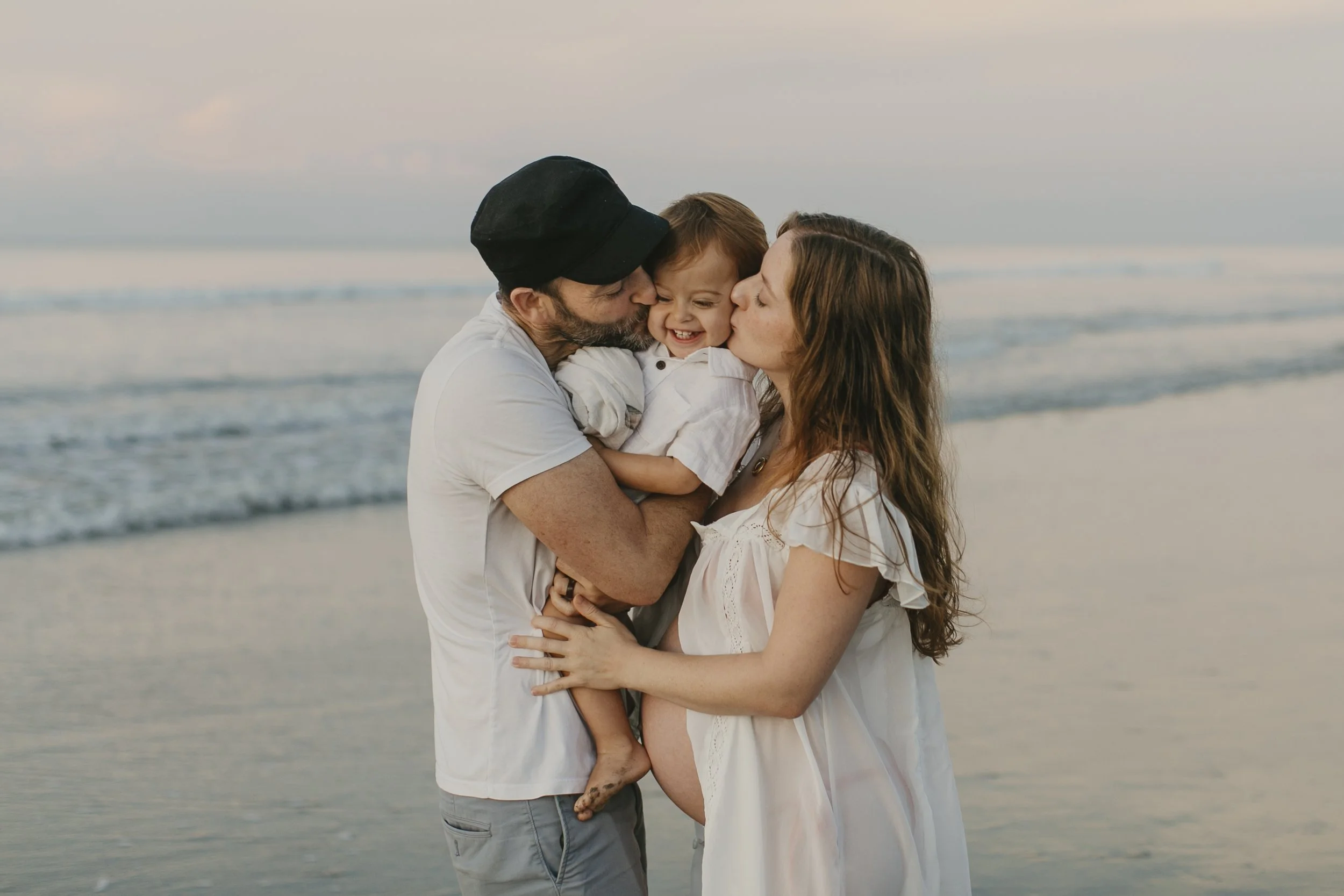 A family of three, a man with a beard and a woman, both embracing their young child at the beach during sunset.