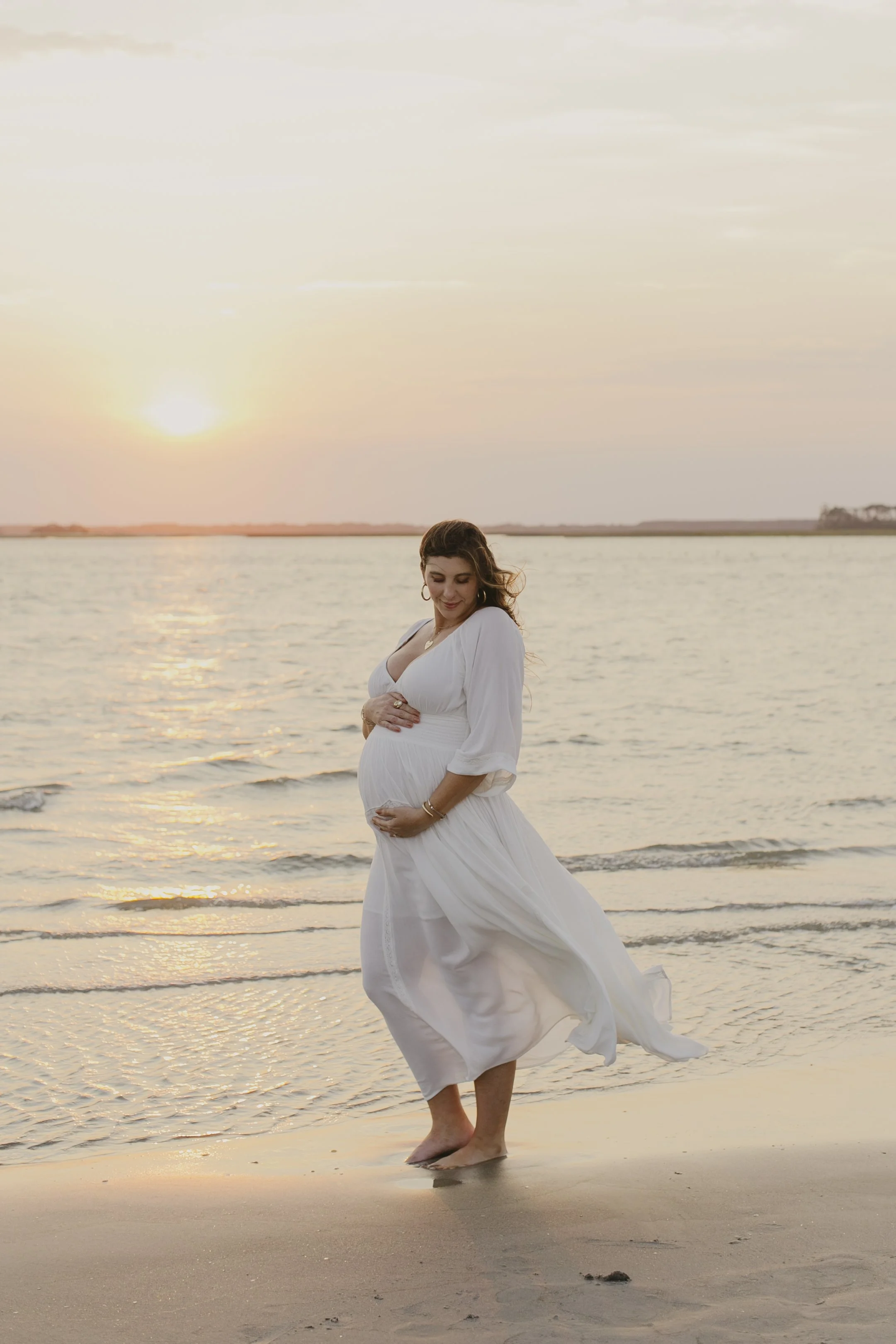A pregnant woman in a white dress walking barefoot on the beach during sunset, with her eyes closed and a peaceful expression.