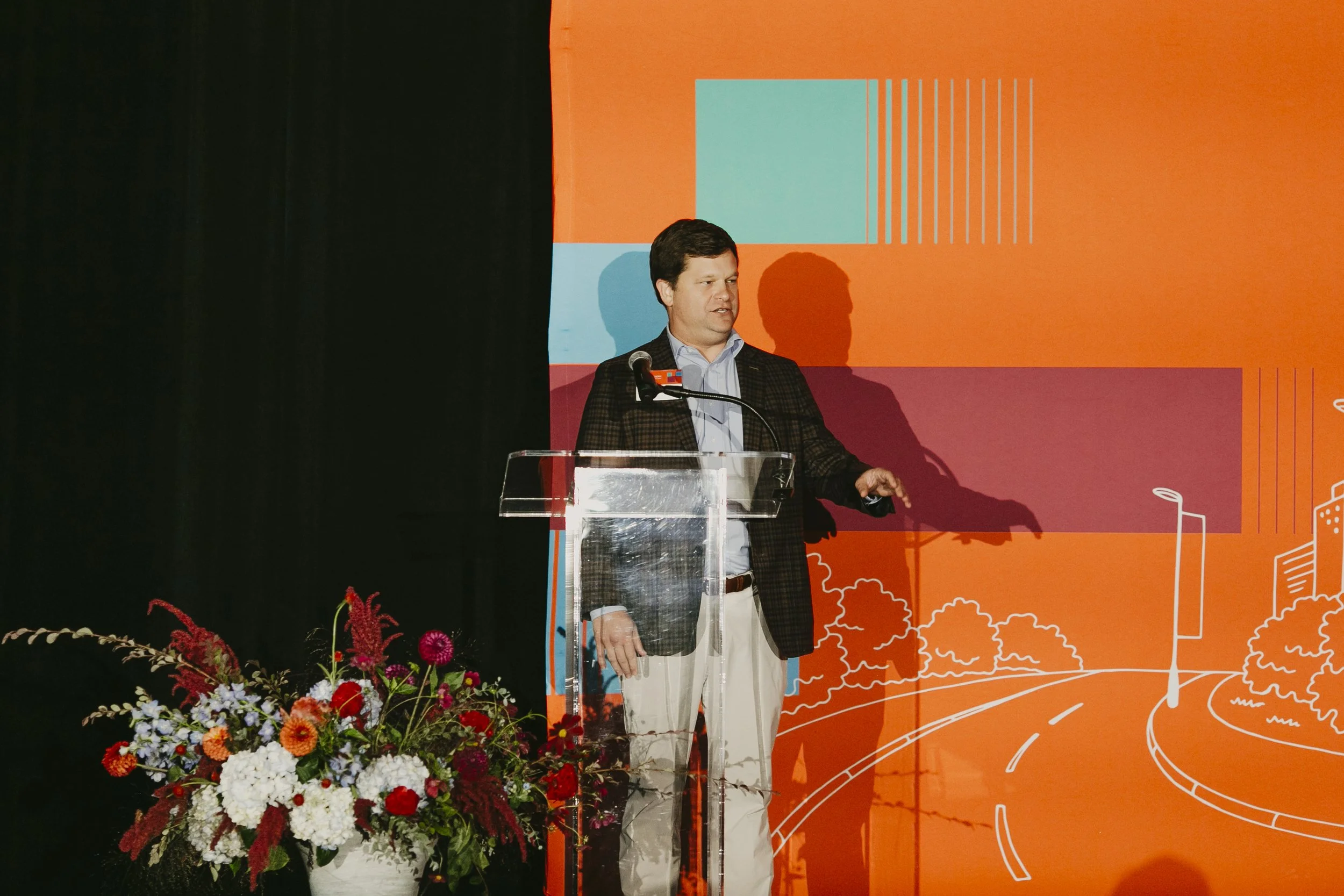 Event emcee speaking at a clear podium on the orange Synovus Journeys to Inspire stage