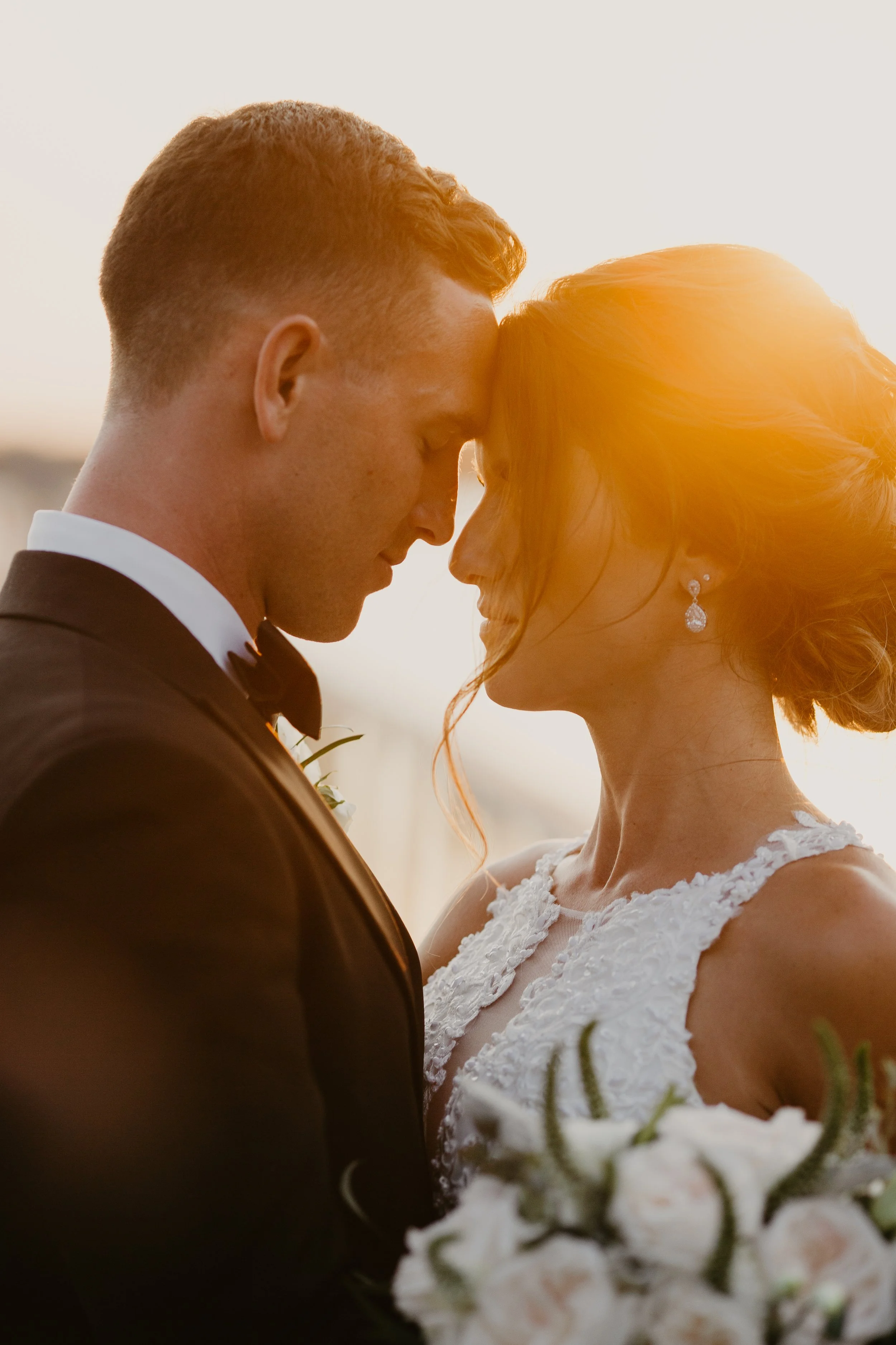 A bride and groom are touching foreheads during their wedding, with a warm sunset glow illuminating their faces. Lowndes Grove Wedding Photos
