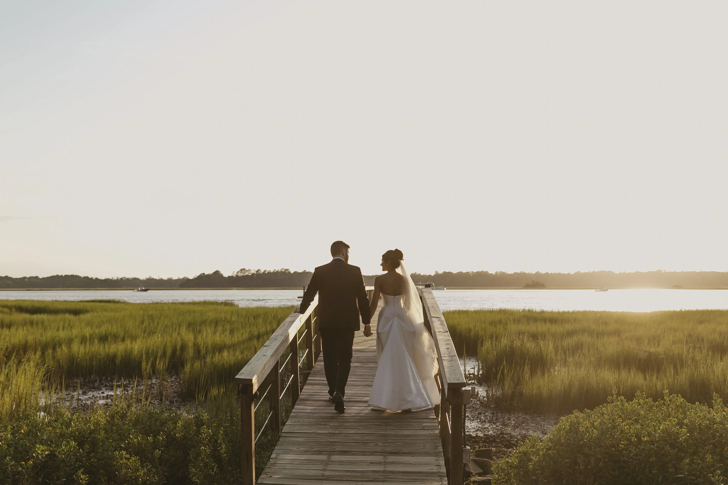A couple dressed in wedding attire walking hand in hand on a wooden dock over a marshland towards a river at sunset.