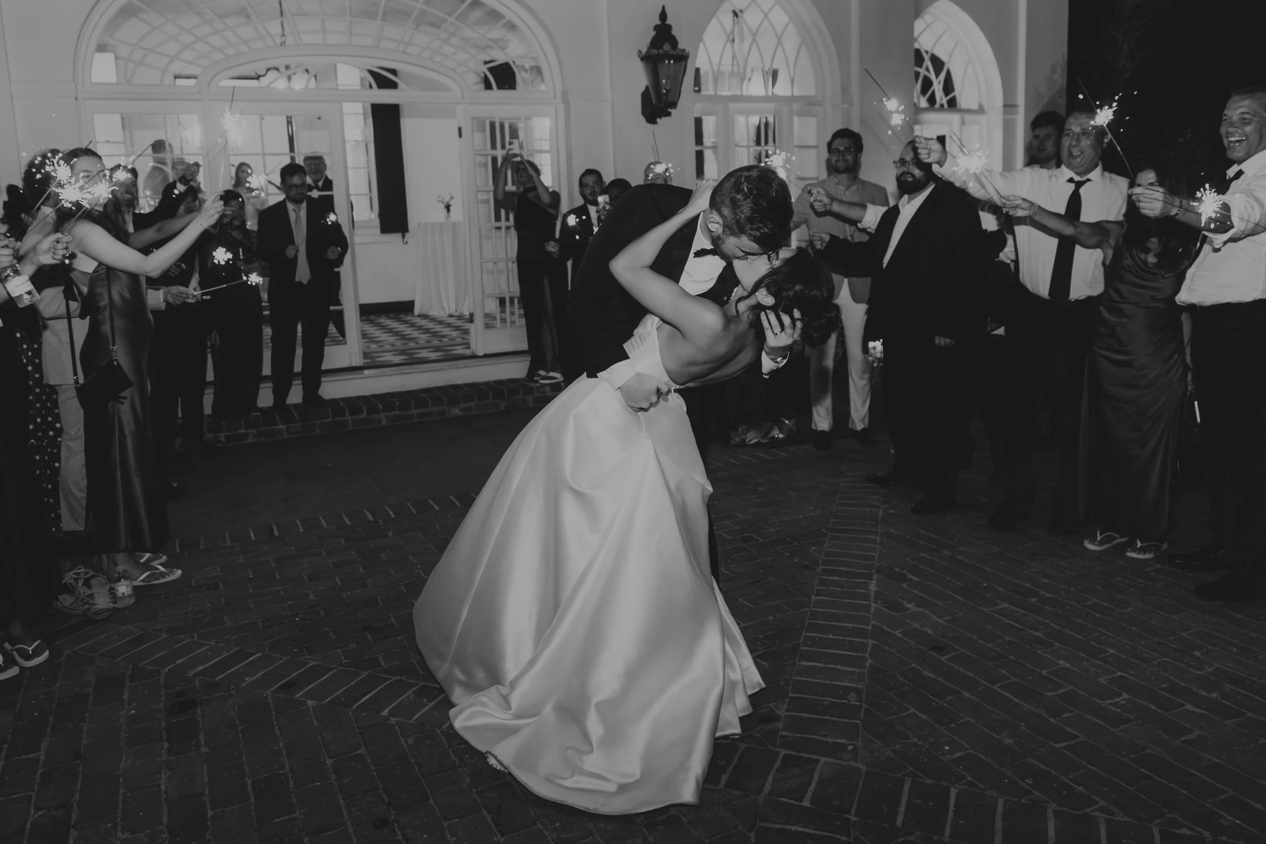 A wedding dance scene where a bride and groom are kissing, surrounded by friends and family holding sparklers. The setting appears to be an elegant indoor venue with large windows and decorative lighting.