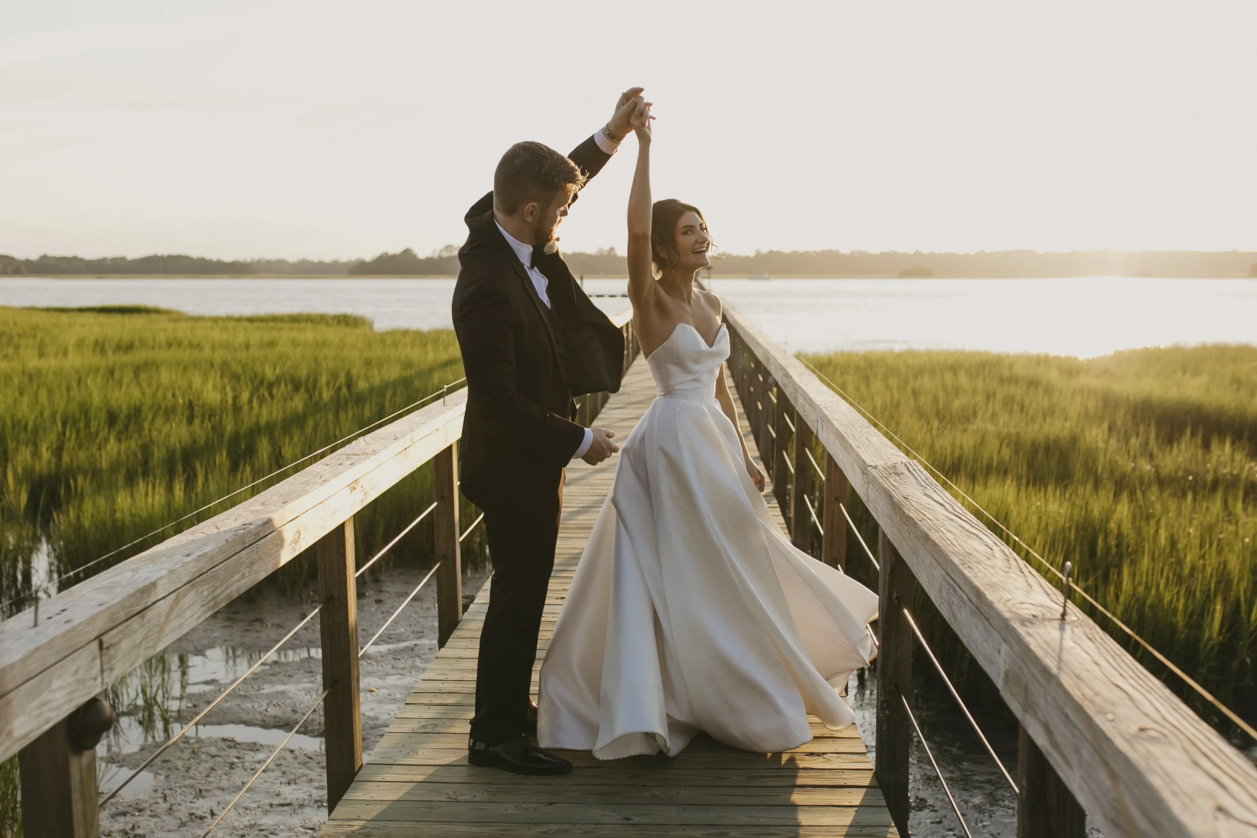 A newlywed couple dancing on a wooden bridge over marshland at sunset, with water and distant trees in the background.