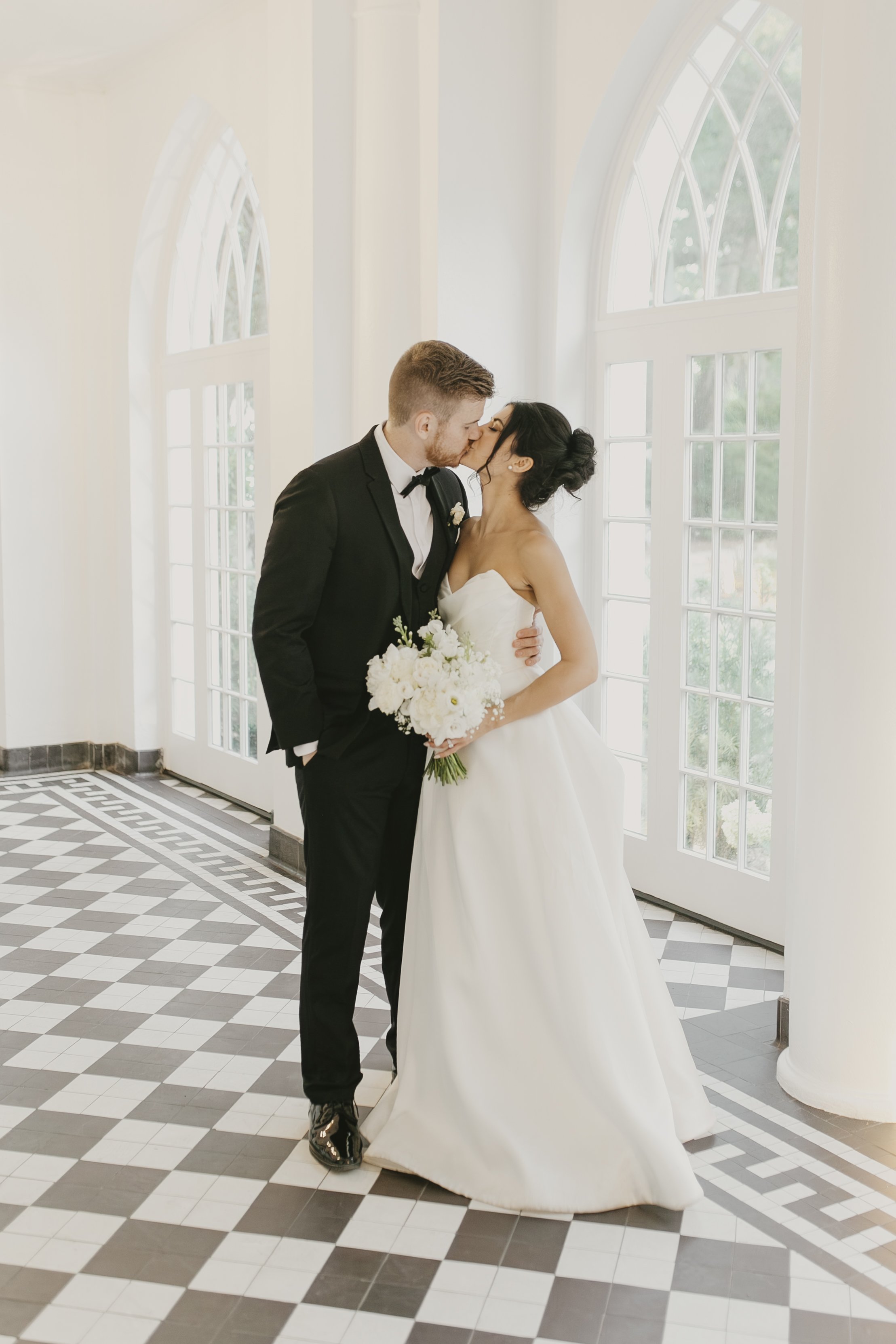 A bride and groom sharing a kiss indoors near large arched windows, the bride holding a bouquet of white flowers, both dressed in wedding attire.
