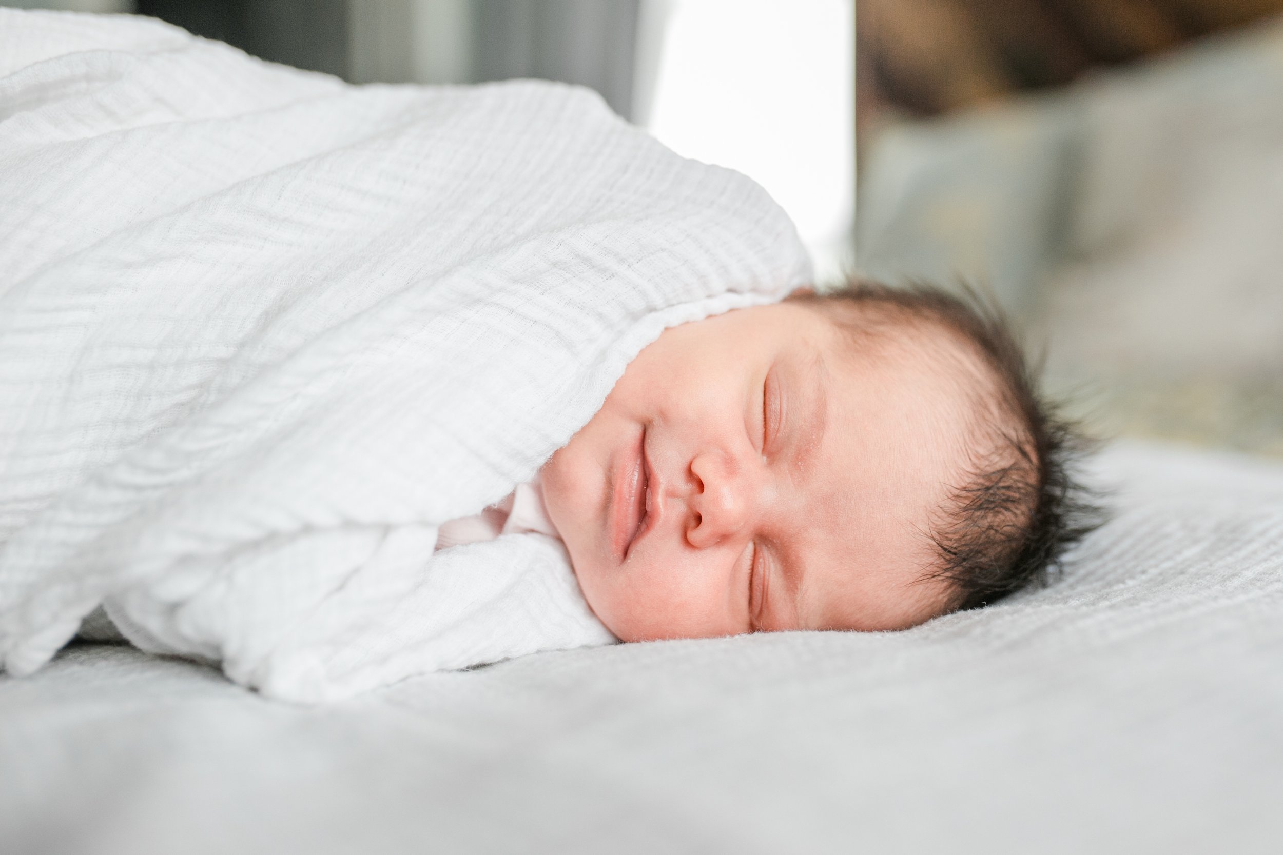 Sleeping newborn baby swaddled in a soft white wrap, smiling during an in-home newborn session in Charleston