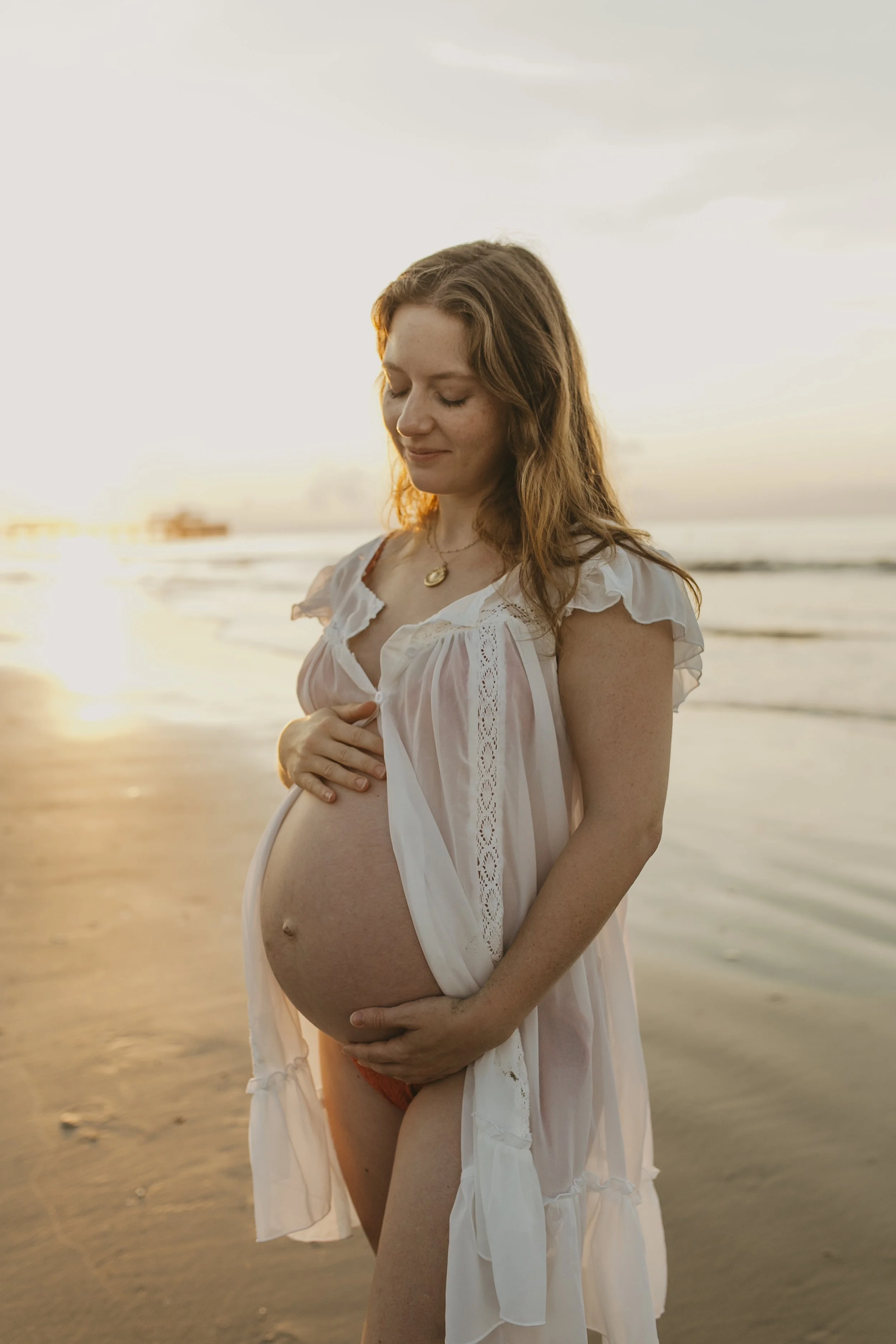 A pregnant woman in a white dress standing on a beach at sunset.