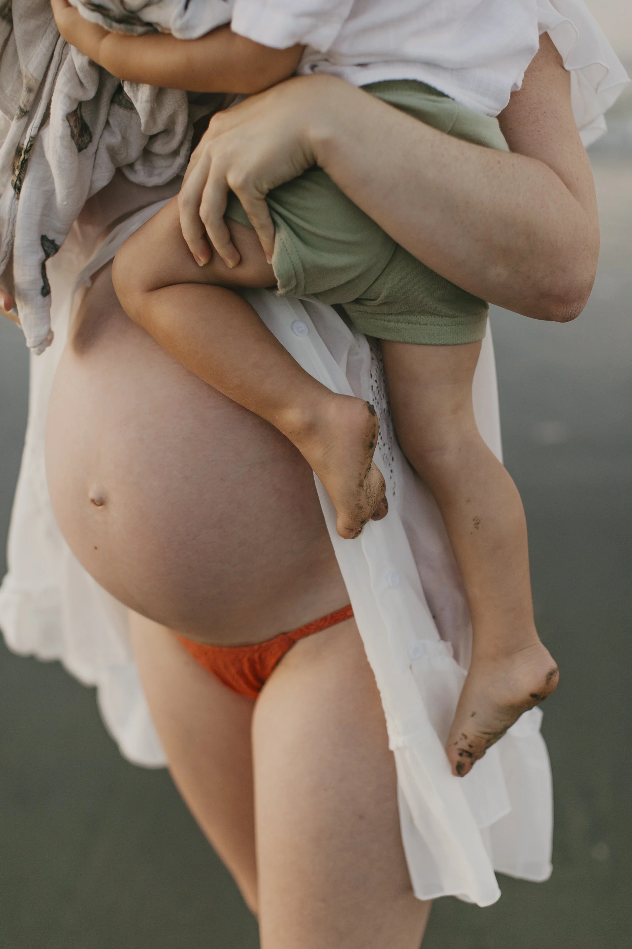 A person wearing a white shirt and orange underwear is holding a small child who is wearing a green shirt and beige shorts. The person has muddy hands and legs and is standing on a gravel or dirt surface.