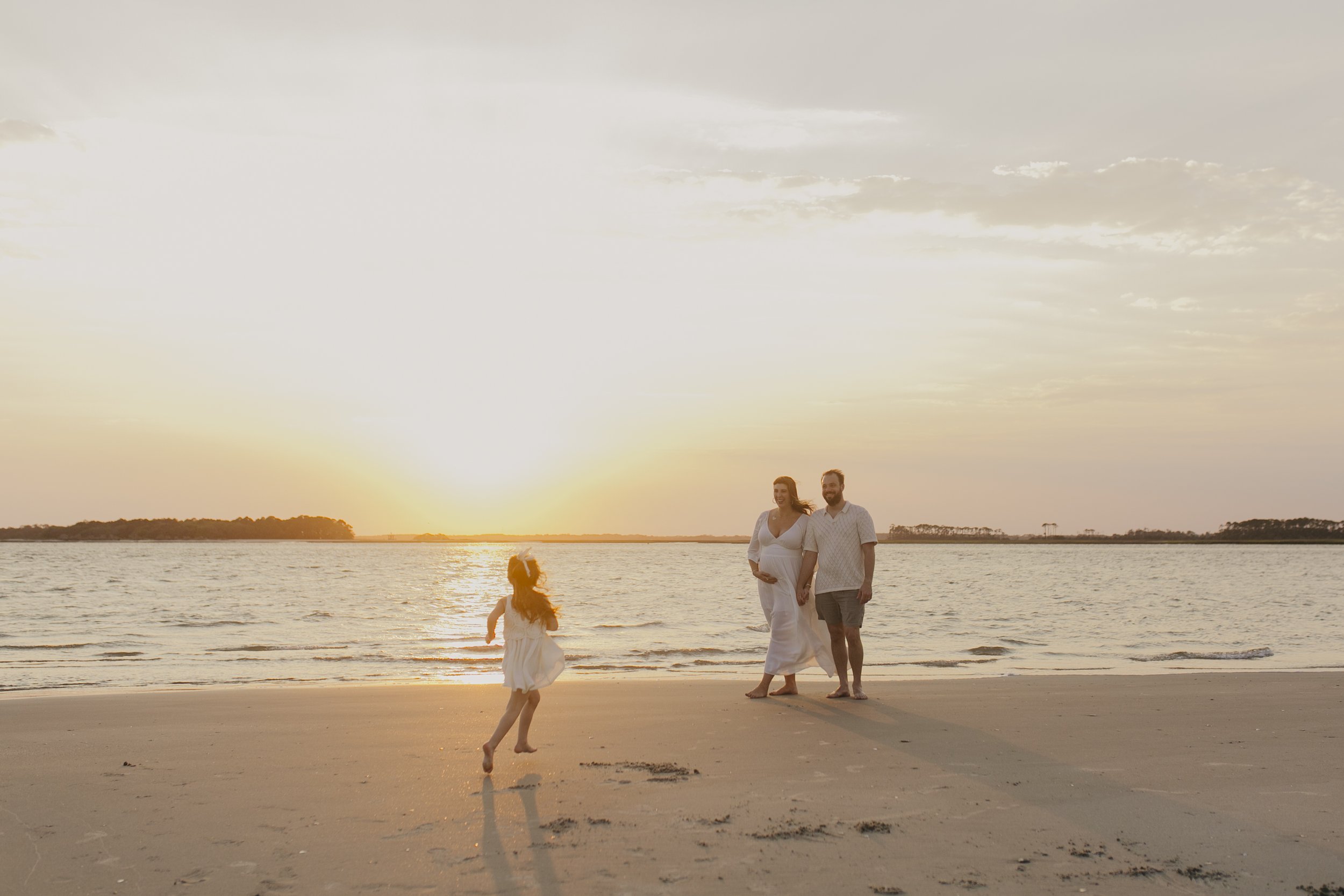 A family enjoying a walk on the beach during sunset, with a woman in a white dress, a man, and a child playing in the sand.