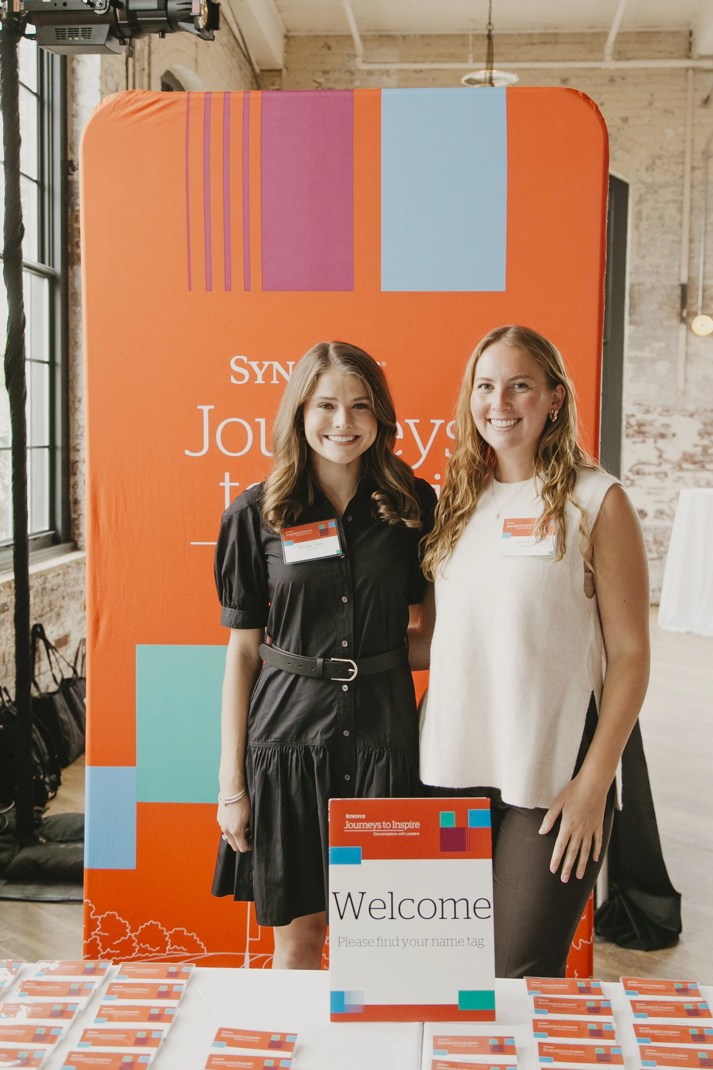 Two event hosts welcoming guests in front of the Synovus Journeys to Inspire backdrop