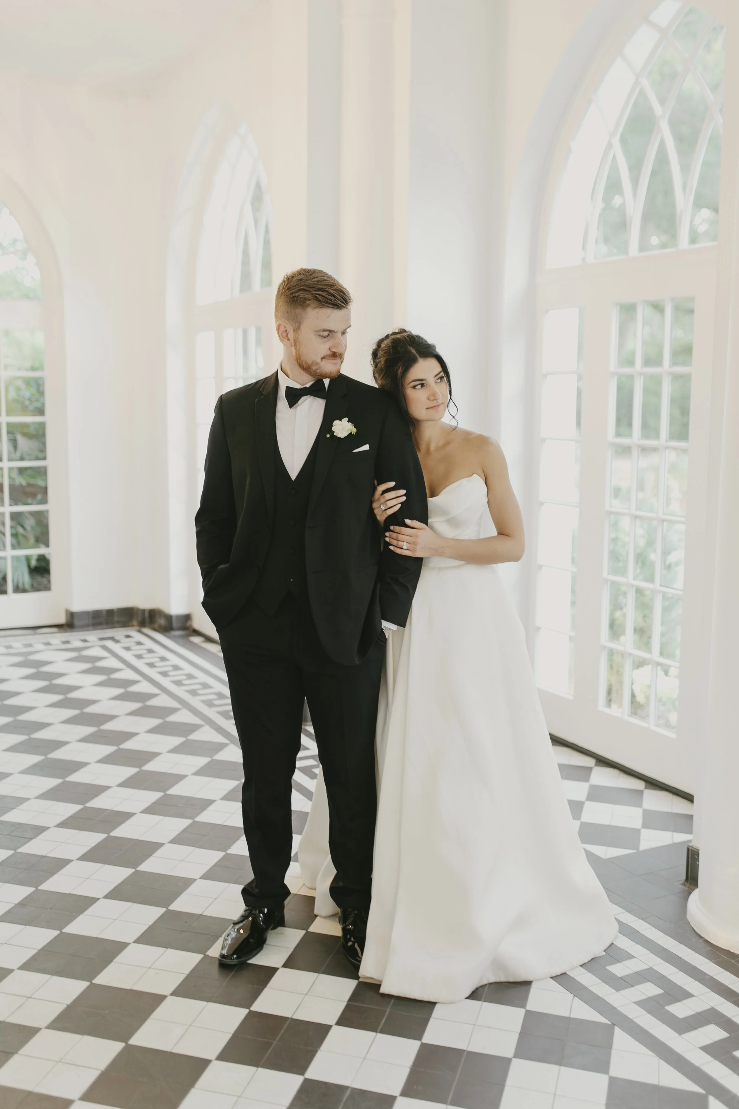A bride in a strapless white wedding gown with her arm linked with a groom in a black tuxedo with a white shirt, black bow tie, and boutonniere, standing in a bright room with large windows and black-and-white checkered floor.