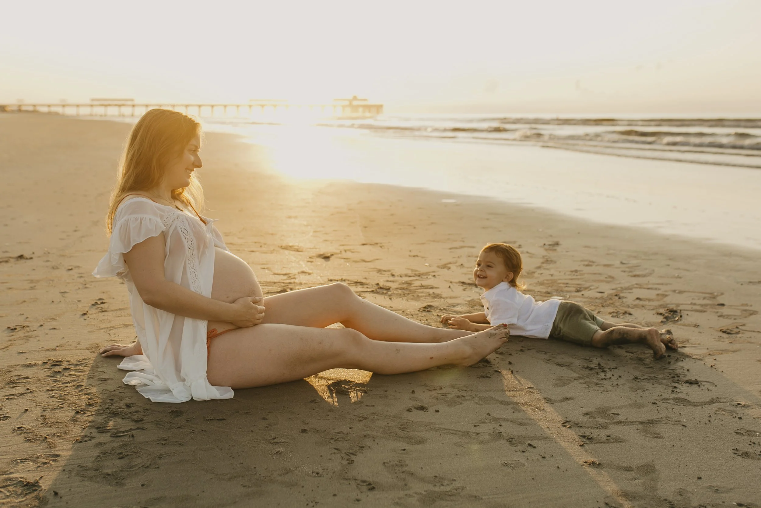 A pregnant woman and a young child sitting and lying on the beach at sunset, smiling and enjoying each other's company.