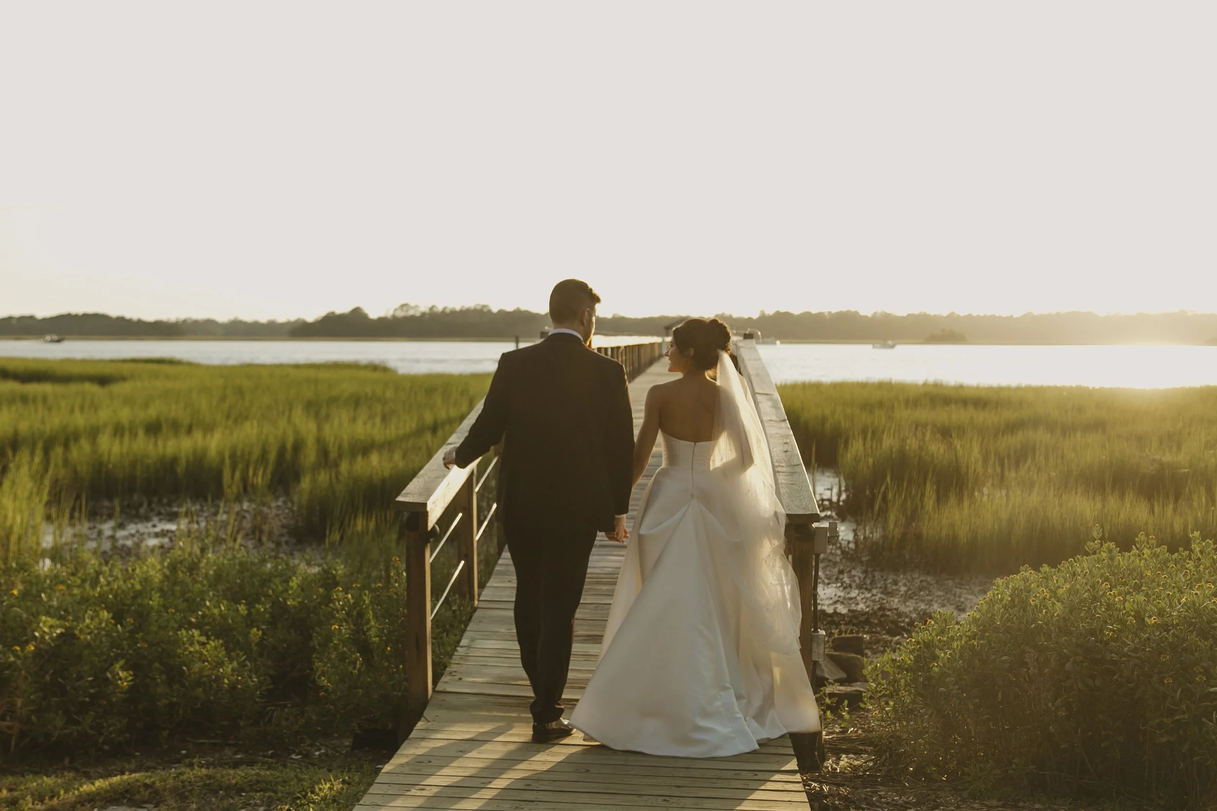 A bride and groom walking hand in hand on a wooden bridge over a marsh at sunset.