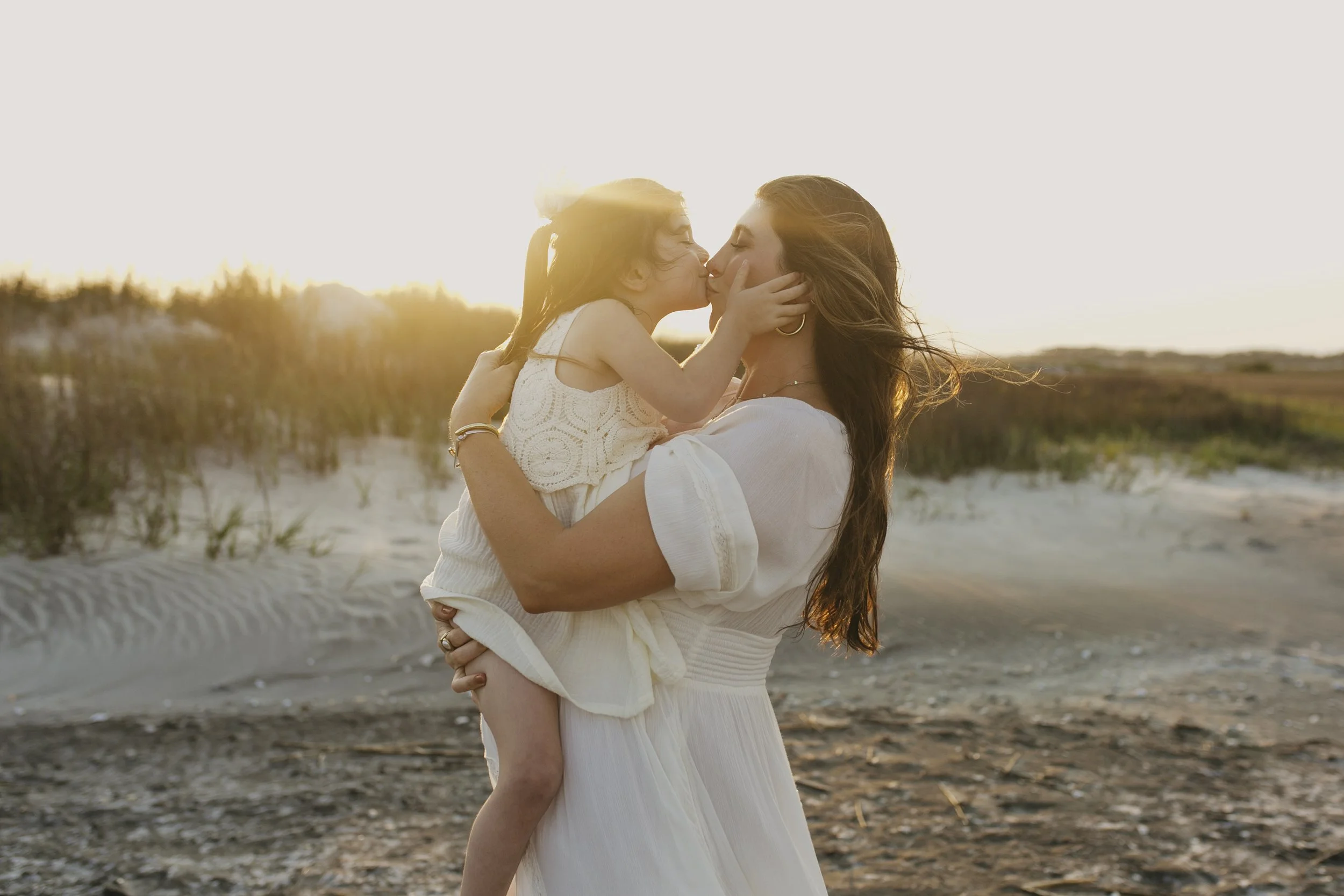 A woman holding a young girl in her arms on a beach during sunset, sharing a kiss.